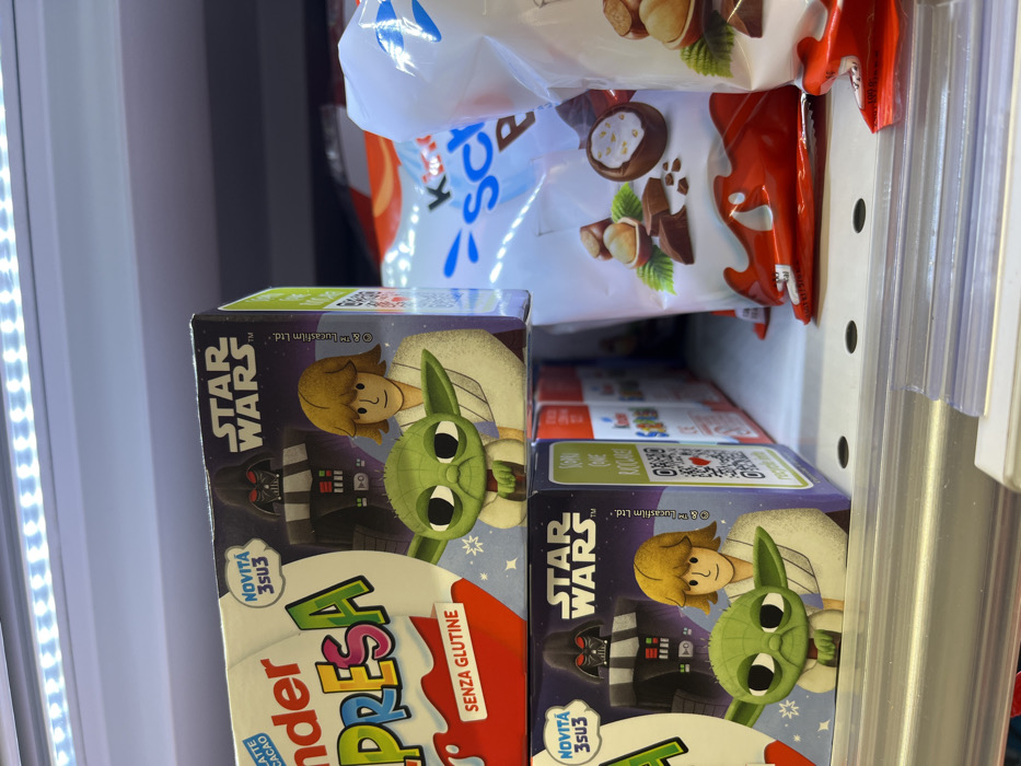 The image displays a retail shelf inside a commercial establishment in Monsummano Terme, Italy. The primary subjects are two stacked Kinder Sorpresa chocolate egg boxes, featuring "STAR WARS" branding. Each box prominently illustrates Darth Vader, Luke Skywalker, and Grogu (Baby Yoda). Visible text on the packaging includes "Kinder Sorpresa," "NOVITÀ 3su3" (New 3-in-3), and "SENZA GLUTINE" (Gluten-free). To the right, multiple white bags containing other Kinder chocolate products, depicting hazelnuts and chocolate pieces, are visible but out of focus. The scene is illuminated by a linear white LED light strip positioned above the shelf. A clear plastic shelf-edge label holder runs horizontally across the bottom of the frame. No individuals are present in the image.