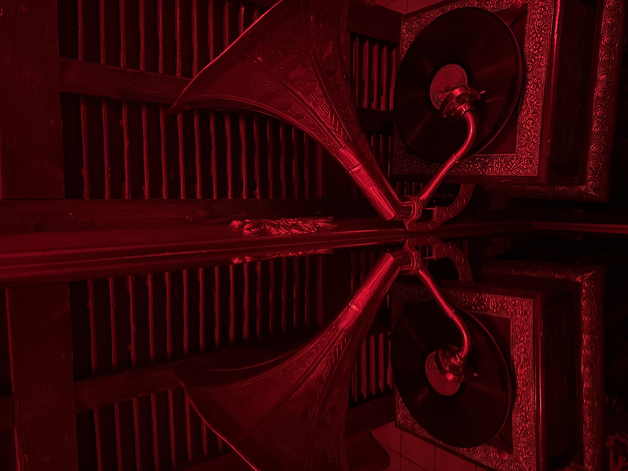 The image shows a vintage gramophone reflecting in a polished surface. The gramophone is predominantly red, with ornate detailing on its horn and base. A vinyl record sits on the turntable. The background features a wooden structure with vertical slats. The reflection mirrors the gramophone and the surrounding environment, creating a symmetrical composition. The image lacks people, actions, or explicit interactions. The location within Tel-Aviv, Israel, cannot be determined from the image alone.