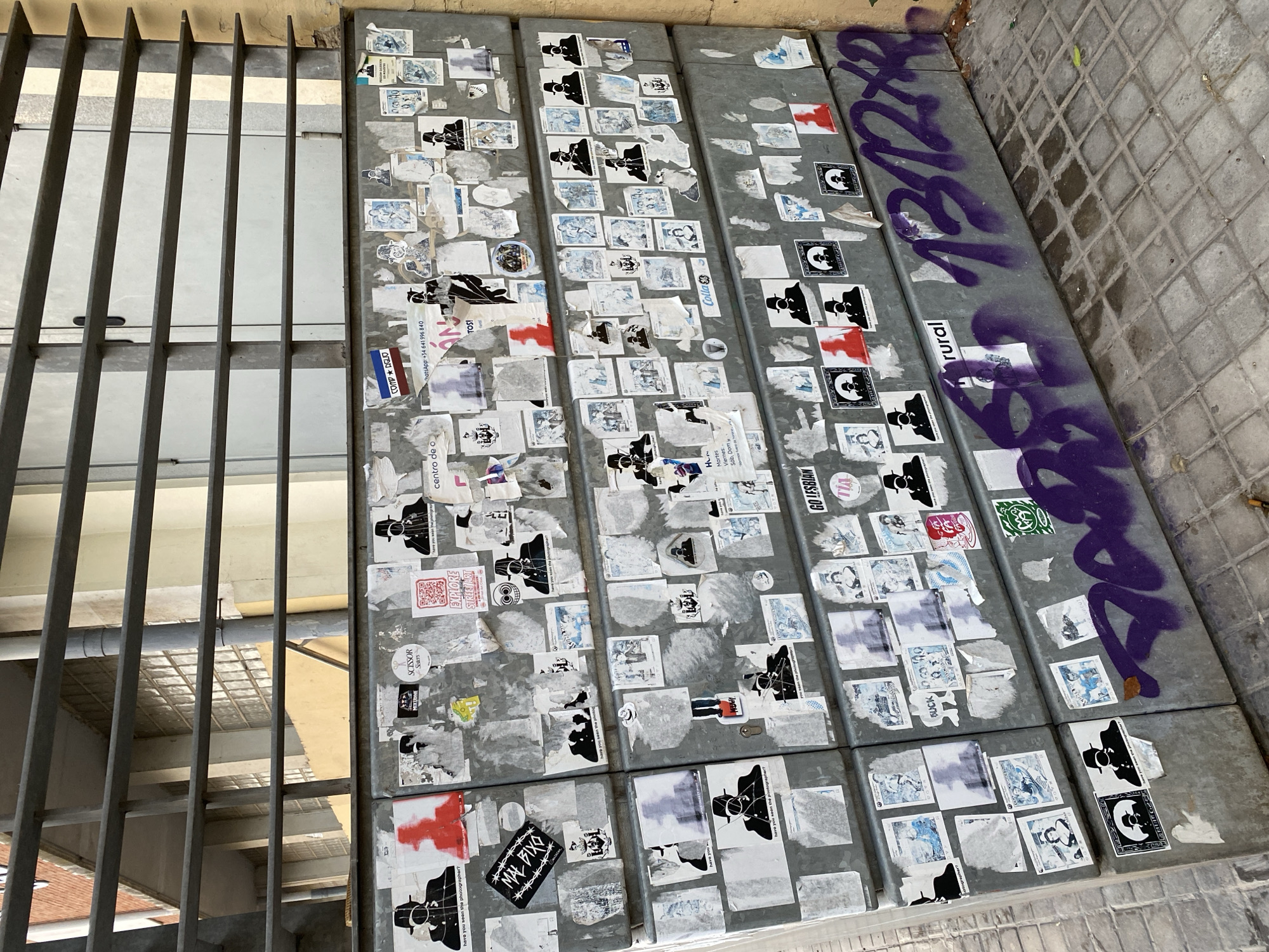 The image shows a close-up view of a graffiti-covered concrete barrier on a city sidewalk. The barrier is located in Valencia, Spain, and is part of a larger structure that appears to be a public seating area or wall. The concrete is gray and weathered, with a textured surface. It is covered in a dense collage of stickers, posters, and graffiti. Many of the stickers feature black and white silhouettes of a person wearing a hat and suit, often with the text "have you seen the photographer?" beneath them. There are also various other stickers with different graphics, including a red silhouette of a person, a QR code, and text in Spanish. The graffiti on the lower portion of the barrier is in purple spray paint and appears to be a combination of numbers and letters, including "1312XR." The sidewalk in front of the barrier is paved with square gray tiles. The background shows a metal grate structure and a building with yellow and white walls. The overall impression is one of urban decay and street art.