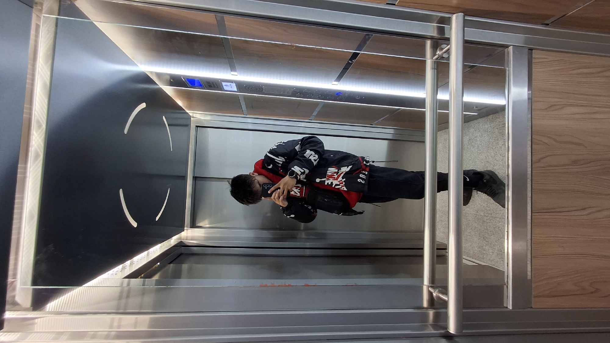 A person is taking a selfie inside an elevator. The elevator has a metal frame and transparent glass doors. The back wall of the elevator is black with four semi-circular, white-colored markings. The ceiling is wood-paneled with a built-in light strip. The person is wearing a black jacket with red accents, black pants, and sneakers. They are holding a smartphone in their hands and appear to be capturing their reflection. The floor of the elevator is gray and appears to be made of tile. The image was likely taken in Varna, Bulgaria, as indicated in the prompt.