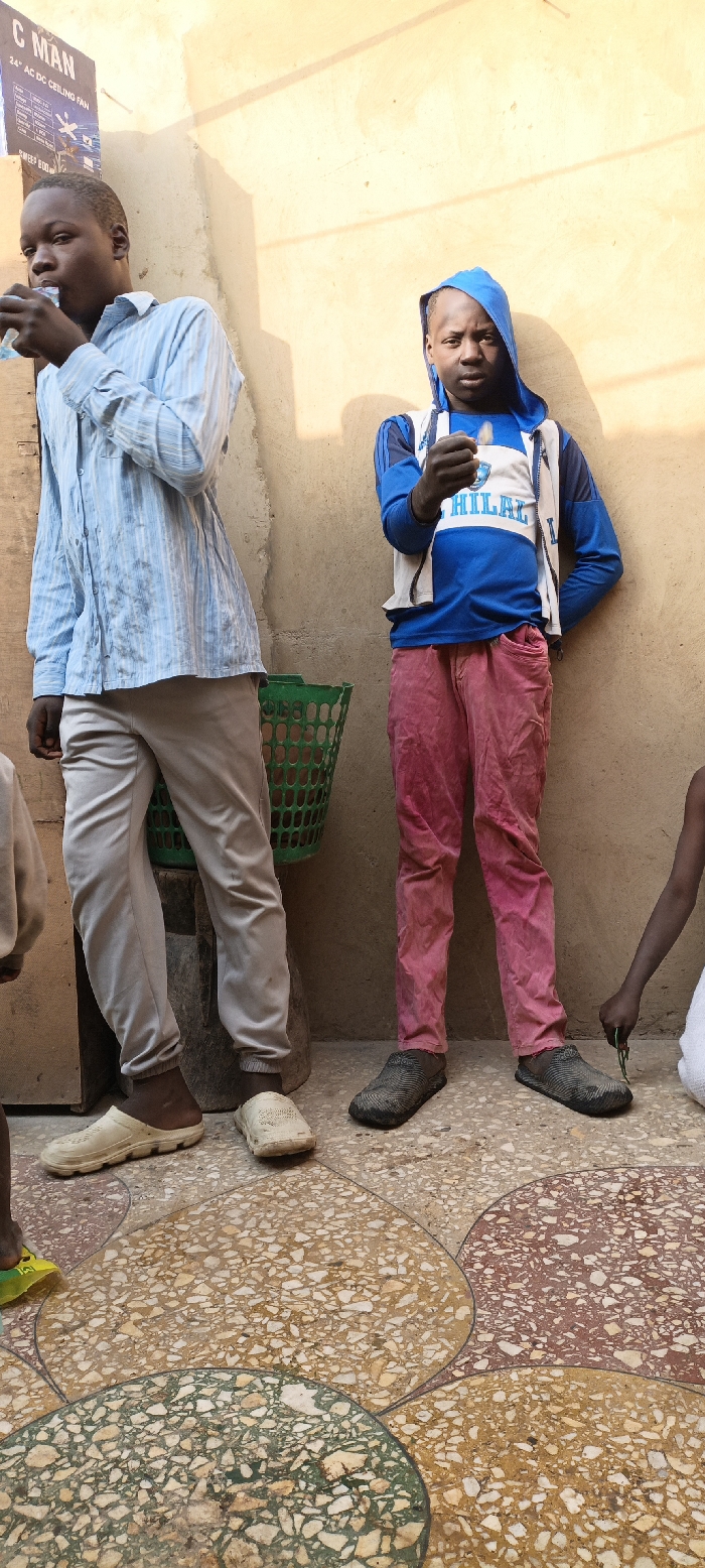A full-color, vertical photograph depicts two young males and a partial third figure standing against a light-colored wall in Kaura Goje, Nigeria. The setting appears to be an outdoor or semi-outdoor area, illuminated by bright daylight, as indicated by the prominent shadows cast on the wall.
On the left, a young man, likely in his late teens or early twenties, wears a light blue striped long-sleeved shirt and light grey trousers. He is captured in the act of drinking from a small, clear plastic pouch or bottle. His feet are clad in off-white clogs.
To his right stands a younger male, presumably a pre-teen or early teen. He is dressed in a blue and white hooded jacket, which features the word "HILAL" prominently on the front. He wears vibrant pink or red pants and dark, textured slip-on shoes. He holds a small, light-colored object in his right hand, looking down towards it.
A partial view of a third, smaller figure, a child, is visible on the far right, wearing a white garment and holding a small green object. Behind the two main figures, a green plastic basket is visible.
On the upper left side of the image, a sign is affixed to the wall. Visible text on the sign includes "C MAN" at the top, followed by "24" AC DC CEILING FAN," and partially visible text indicating "SWEEP 800MM."
The ground features a distinctive terrazzo floor patterned with large, irregular shapes in shades of light brown, green, and reddish-brown, separated by dark lines. The scene conveys a casual, everyday moment in a local setting.