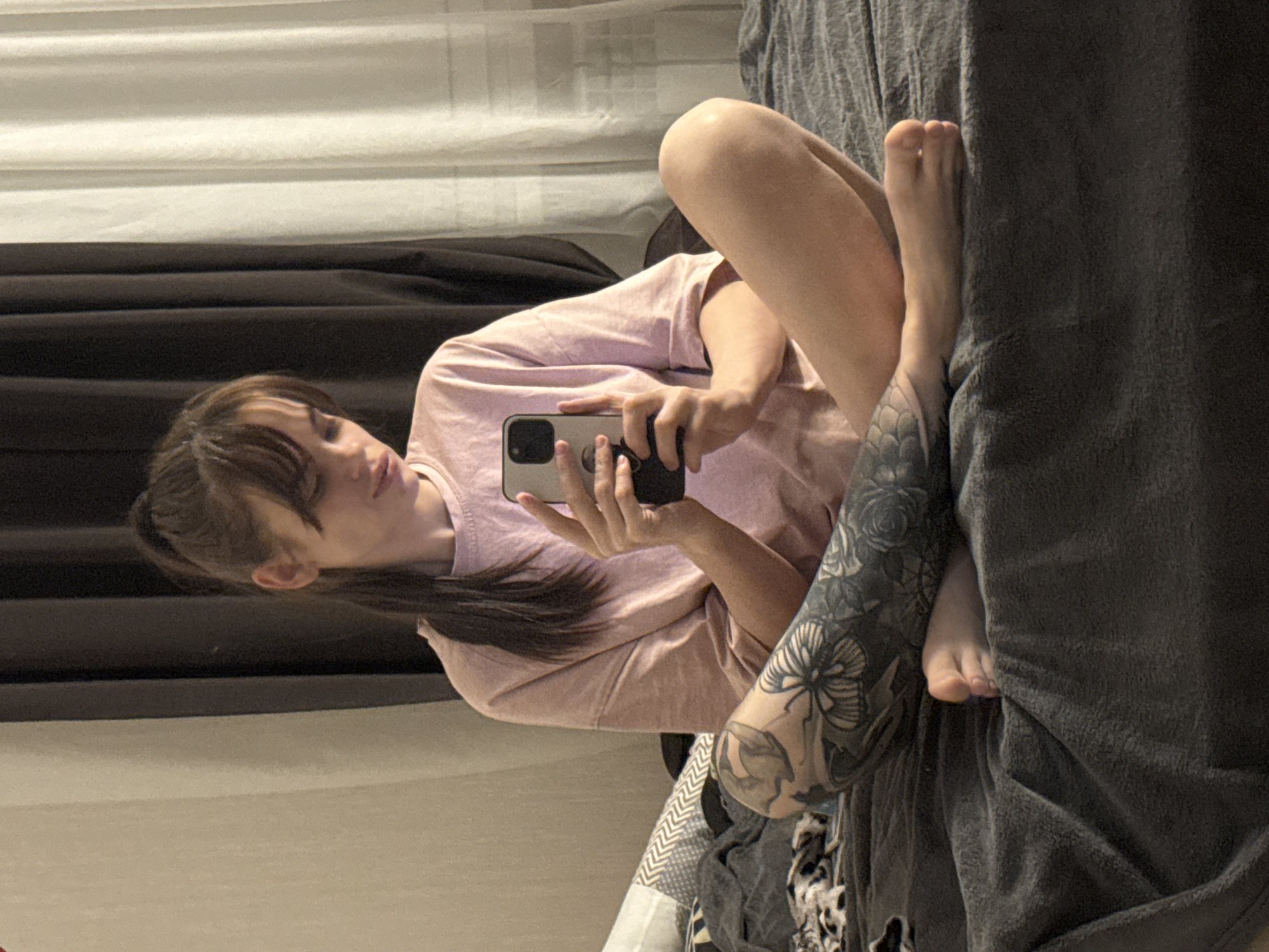 A young woman is sitting on a bed, facing the camera and holding a smartphone. She is wearing a light pink short-sleeved shirt. Her hair is dark and pulled back in a ponytail. Her left leg is extended and features a large floral tattoo. A gray blanket is partially covering the foot of her left leg. The background includes light and dark curtains. There is no indication that this image was taken in Rostov-on-Don, Russia.