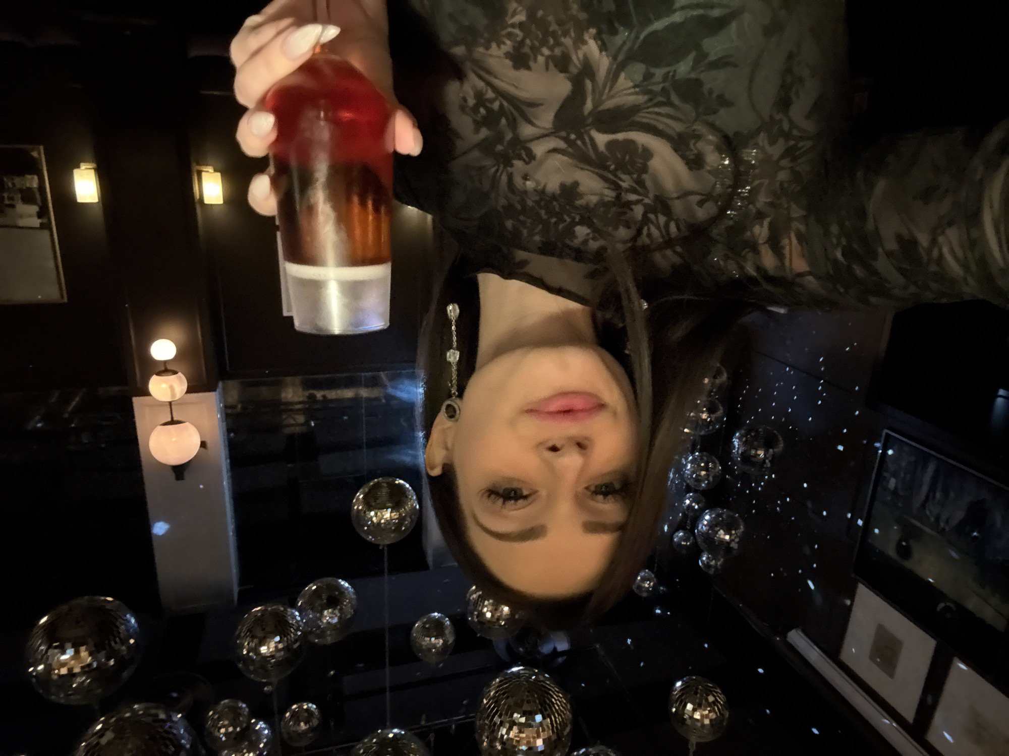 A woman is holding a cocktail in a bar, likely in Moscow, Russia. She is wearing a sheer, dark floral top and dangling earrings. The cocktail appears to be multi-layered, with red, amber, and a light-colored base. The bar has a dark interior, with disco balls hanging from the ceiling and framed artwork on the wall. There are sconce lights and globe lights visible in the background. The image is oriented upside down.