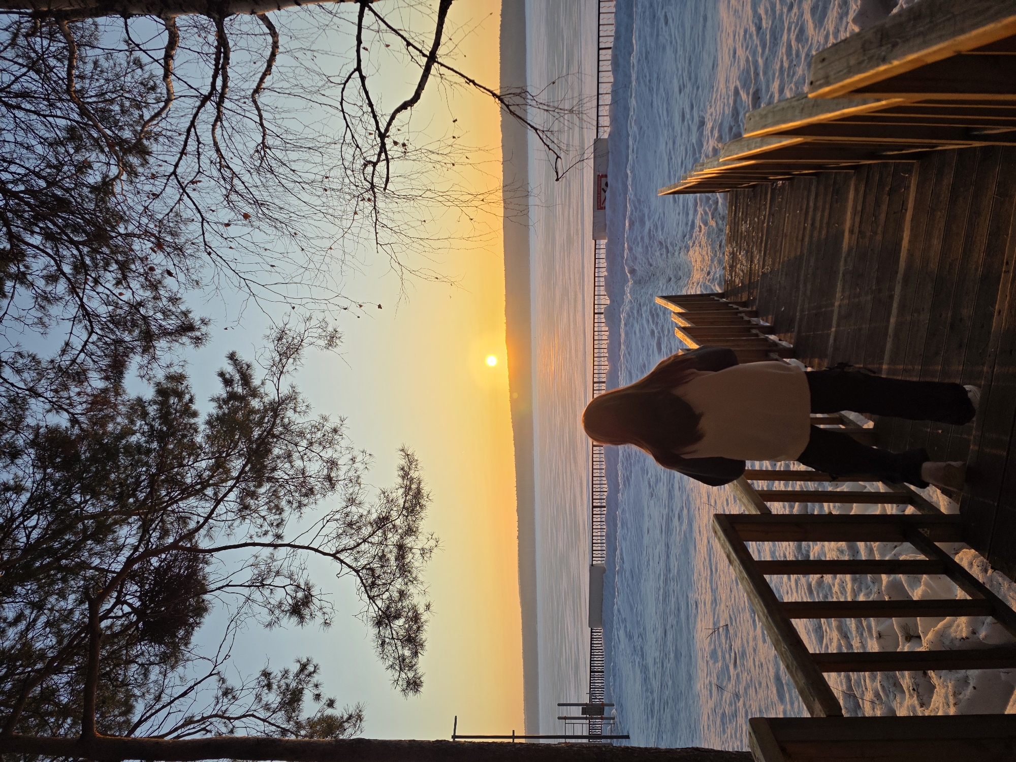 The image depicts an outdoor winter scene at either sunrise or sunset over the Votkinsk Reservoir, located in Chaikovsky, Russia. In the foreground, a person with long brown hair, wearing a light-colored top, dark outer layer, and dark pants, is observed from behind, descending a wooden staircase. The staircase has wooden railings on both sides, leading down towards a snowy embankment. The ground in the midground is covered in snow, showing some tracks. A black metal fence or railing spans horizontally across the scene, separating the snowy area from the frozen expanse of the reservoir. Behind the fence, a white bench or sign and a metal structure (possibly playground equipment) are partially visible to the left. The Votkinsk Reservoir occupies the background, appearing as a vast, flat, frozen surface. The sun, a bright orange disk, is positioned slightly right of center, just above the reservoir's horizon, casting a warm orange and yellow glow across the sky that transitions to pale blue overhead. A dark, forested line of land is visible on the far horizon. Silhouetted branches of coniferous and deciduous trees frame the upper portion of the scene.