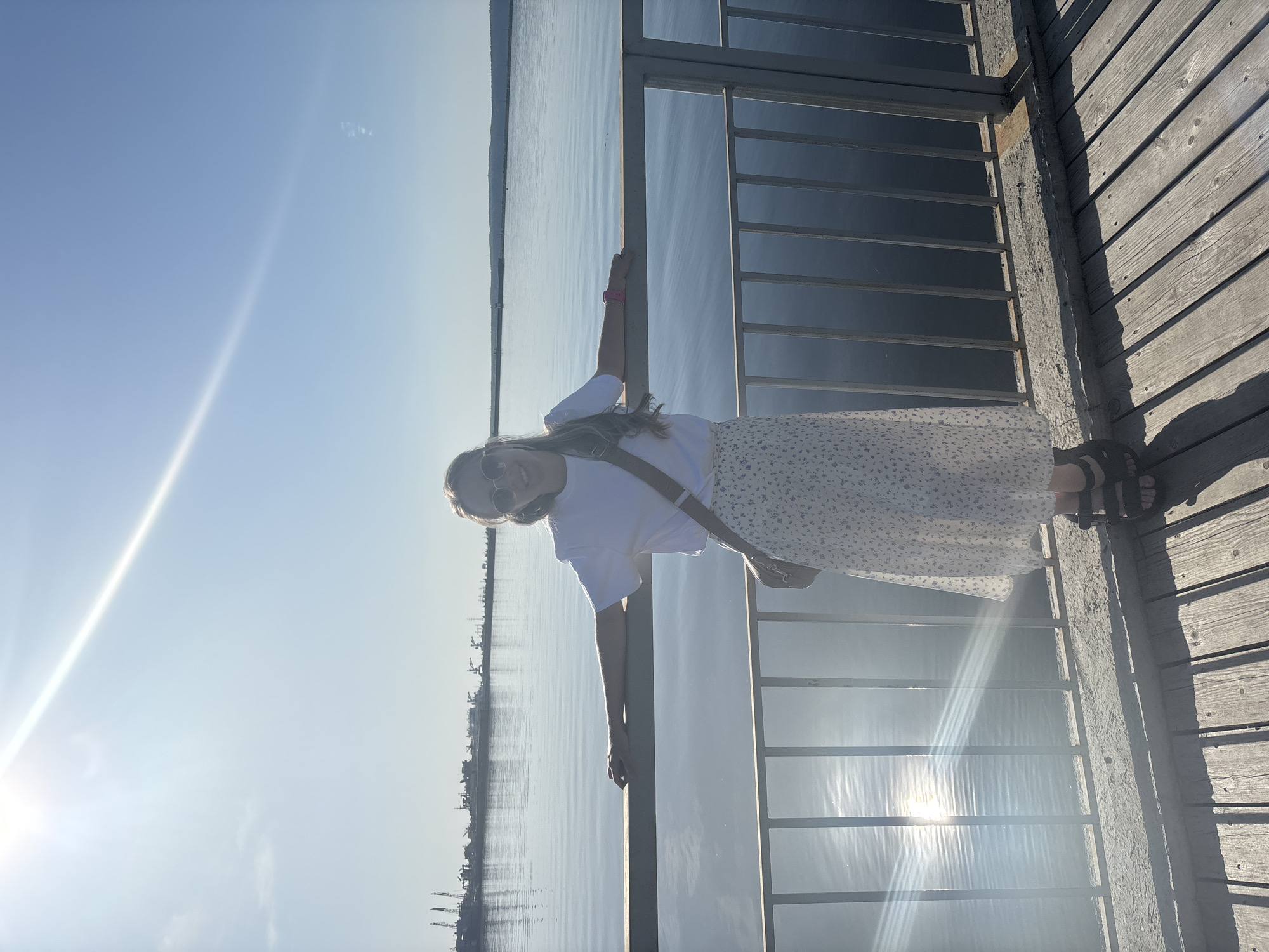 The image shows a person standing on a wooden pier with metal railings, possibly in the city of Chaikovsky, Russia, suggested by the presence of an industrial skyline across the water. The individual, appearing female, wears sunglasses, a white t-shirt, and a light-colored, floral-patterned skirt. She has a strap across her shoulder, likely from a bag. She leans on the railing with one hand. The scene is brightly lit, with the sun low in the sky, causing a glare on the water. The sky is clear and blue.