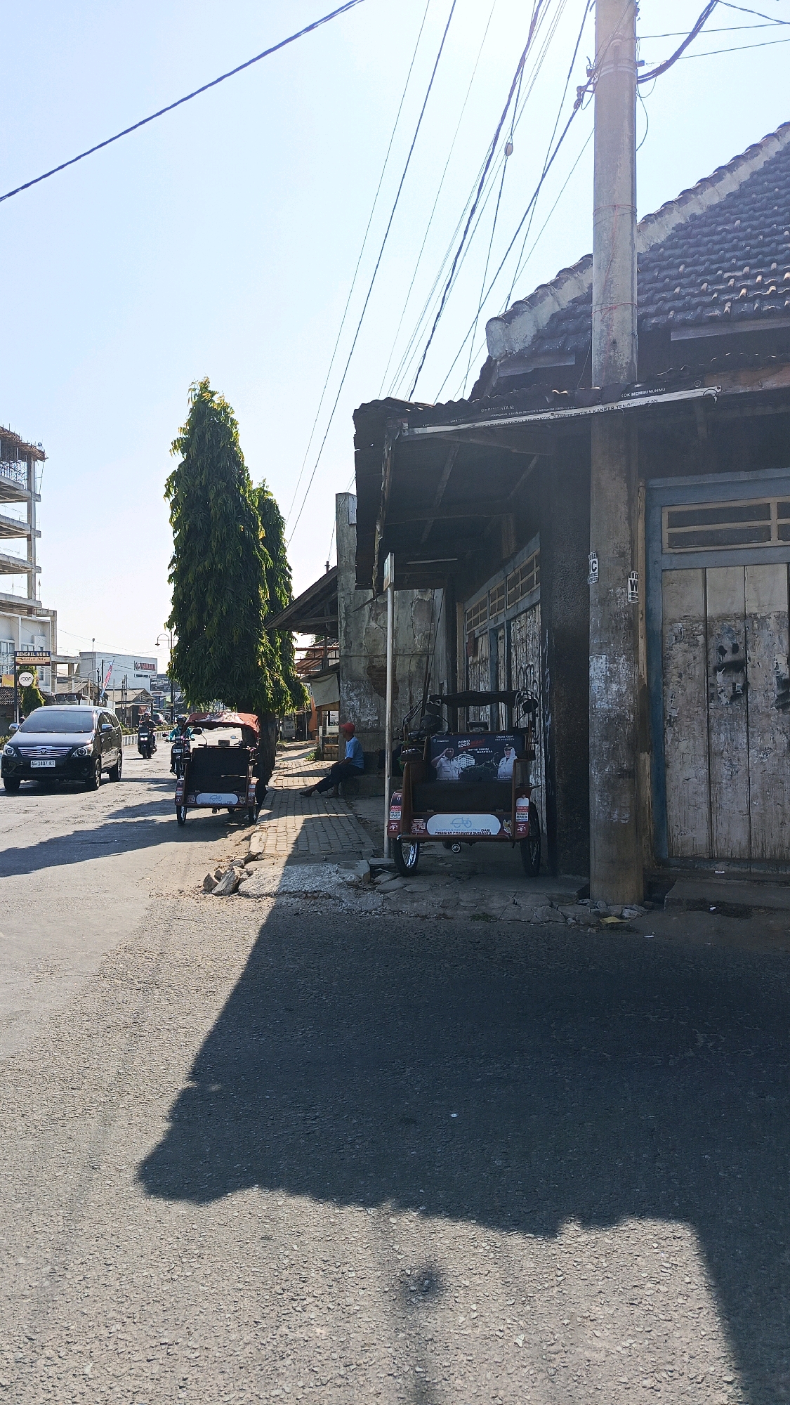 The image depicts a street scene in Kertosari, Indonesia, on a clear, sunny day. In the foreground, the asphalt road casts a long shadow, suggesting it is midday. On the right side of the road, a weathered building with a tiled roof and wooden doors stands. A utility pole with numerous overhead wires is situated next to the building. Parked along the sidewalk in front of the building are two three-wheeled vehicles, possibly autorickshaws, with passengers visible inside the one on the right. On the left side of the road, a black SUV is driving away from the camera, followed by a motorcycle. Further down the street, a tall, slender evergreen tree is planted on the sidewalk, and beyond it, construction of a multi-story building is underway. A person wearing a blue shirt and red cap sits on a ledge near the building. The environment appears to be a mixed urban and commercial area.