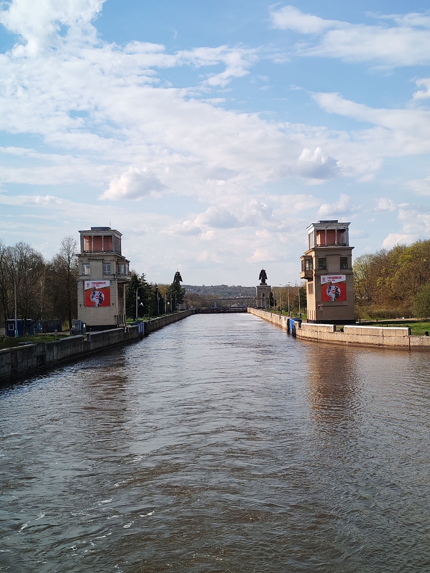 The image shows the Moscow Canal in Yakhroma, Russia. The water is dark and rippled, reflecting the sky and surrounding structures. On either side of the canal are concrete walls and two-story buildings with distinctive red-trimmed roof structures. Each building has a large advertisement poster on its side that reads "ГОРДИМСЯ!" Statues are positioned on either side of the canal, behind the buildings. Trees line the banks of the canal on both sides. In the distance, a bridge crosses the canal. The sky is blue with scattered white clouds.