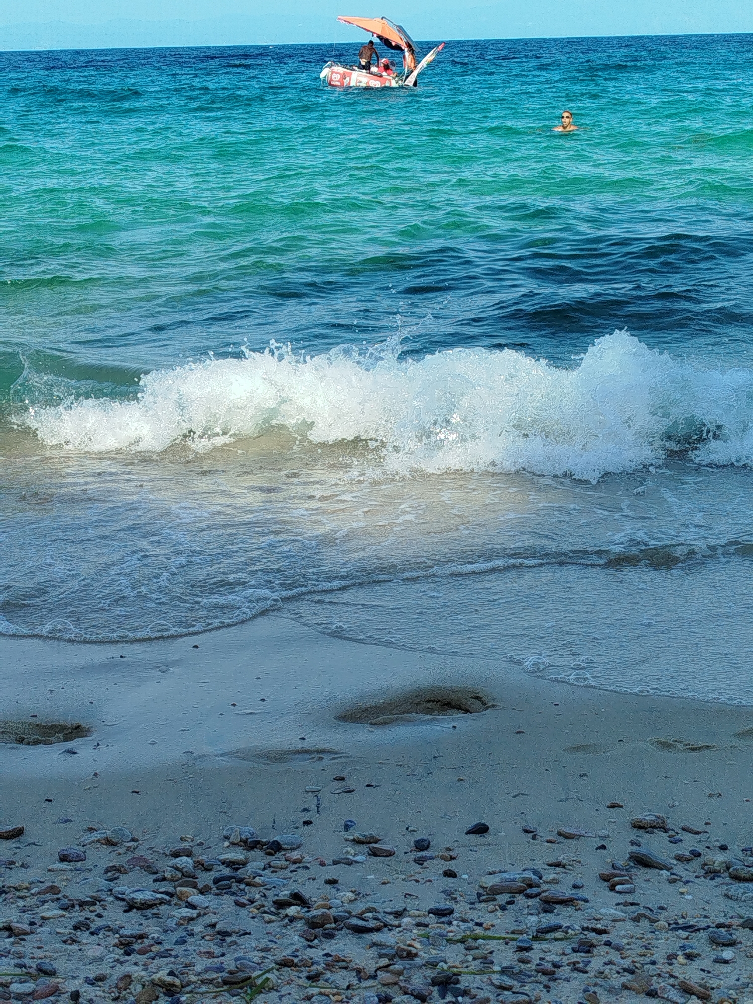 The image depicts a beach scene in the Sithonia Municipal Unit, Greece. In the foreground, a sandy beach is covered with small pebbles and stones. A gentle wave is breaking on the shore, with white foam spreading across the wet sand. The water transitions from a lighter turquoise near the shore to a deeper blue further out. In the middle ground, a person is swimming in the ocean, appearing to wear sunglasses. In the background, a small inflatable raft or boat with an orange canopy is visible, with several individuals on board. The sky is clear and blue.