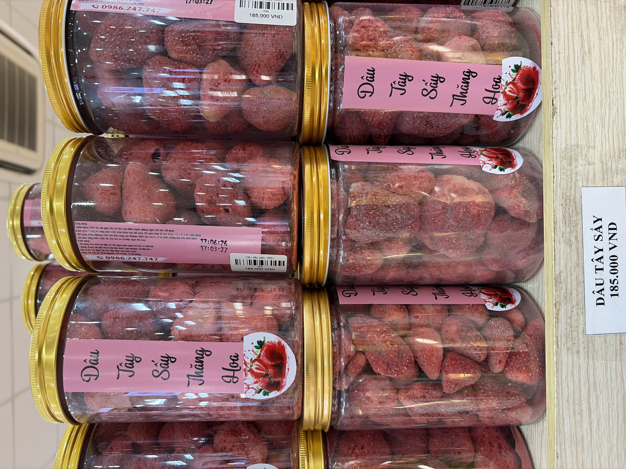 The image displays a retail setting in Đà Nẵng, Vietnam, with a focus on food products. Shelves are stacked with clear plastic jars filled with dried strawberries. The jars have gold-colored lids. Several jars feature pink labels with Vietnamese text, including "Dâu Tây Sấy Thăng Hoa" (Dried Strawberries Blossoming). Some labels include small illustrations of strawberries. The lighting suggests an indoor environment, possibly a grocery store or market. A price tag is visible at the bottom of the image, indicating "DÂU TÂY SẤY 185.000 VNĐ".