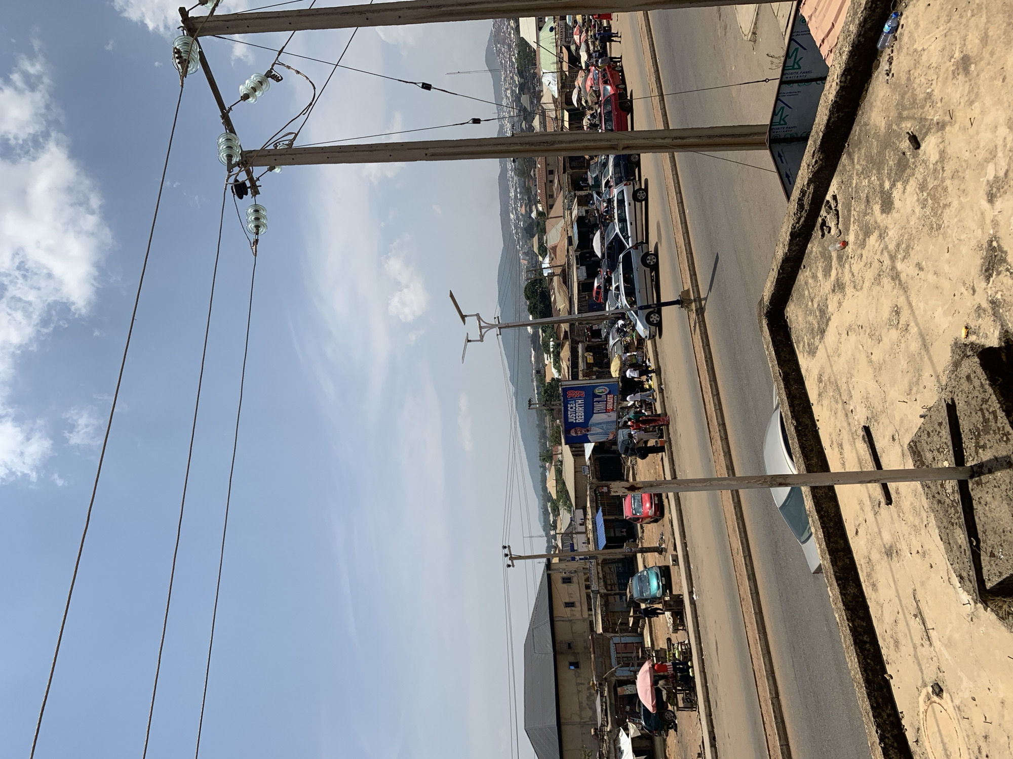 The image shows a street scene in Rafin Par, Nigeria. The view is from an elevated position, possibly a building. A two-lane road runs along the center of the frame, with vehicles parked along one side. Street vendors with carts are present near the parked cars. Buildings of varying heights and conditions line the street. Mountains are visible in the distance. Power lines and utility poles are prominent in the foreground. The sky is partly cloudy. The concrete ledge of the building from which the photo was taken is visible in the right foreground.