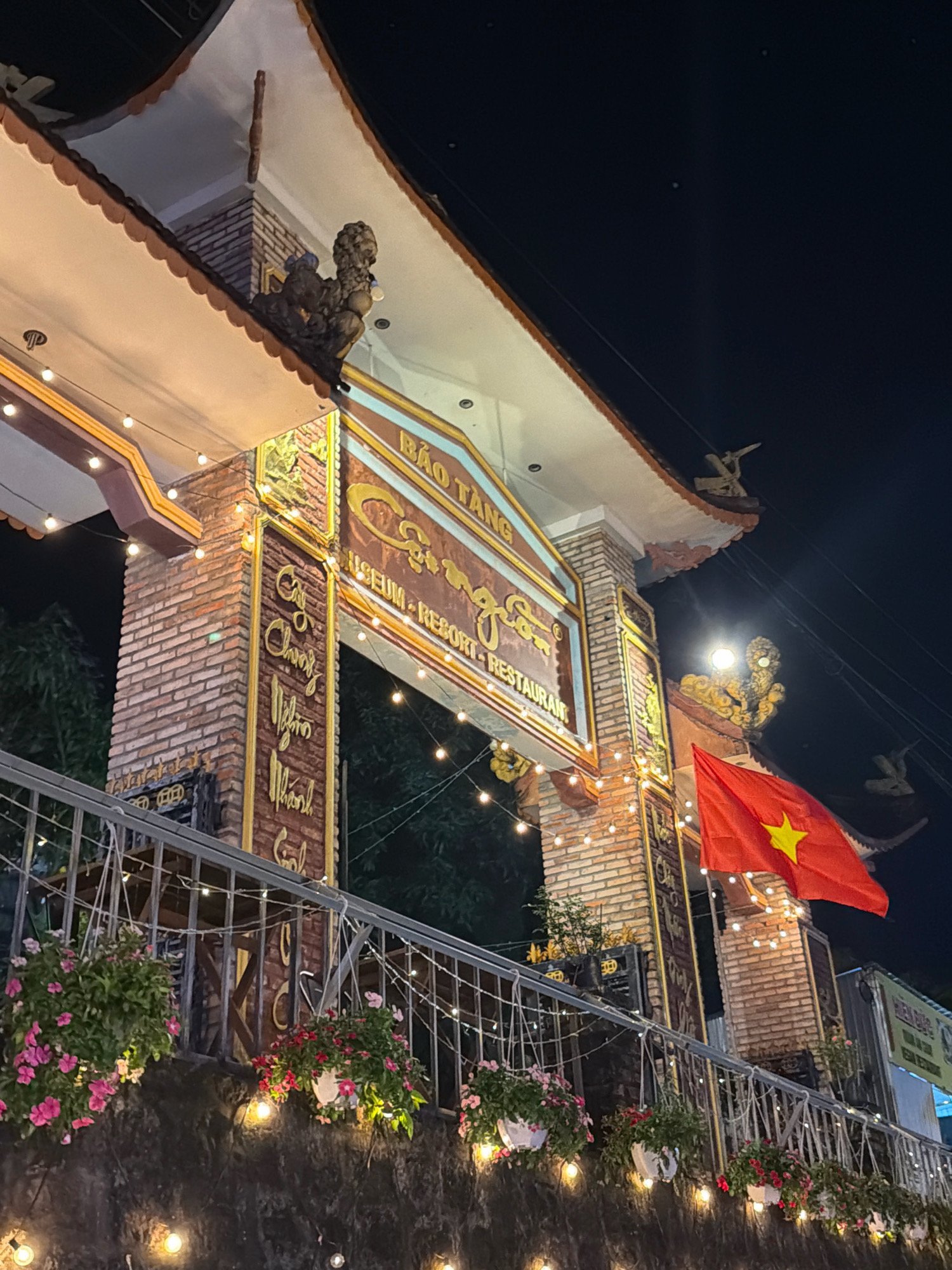 This image captures the entrance of the "Bao Tang Cung Son Museum, Resort & Restaurant" located on the island of Phú Quốc, Vietnam, at night. The architecture features a traditional Vietnamese style with a prominent archway constructed from red brick and adorned with decorative elements. A Vietnamese flag with a yellow star is prominently displayed on the right side of the archway, flying in the night breeze. Strings of warm, glowing lights are strung along the archway and a metal railing in the foreground, illuminating the scene. Hanging baskets filled with pink and red flowers are suspended from the railing, adding a touch of natural beauty to the urban setting. The background is dark, with some trees visible, suggesting the establishment is surrounded by greenery. The overall impression is one of a welcoming and atmospheric nighttime destination.