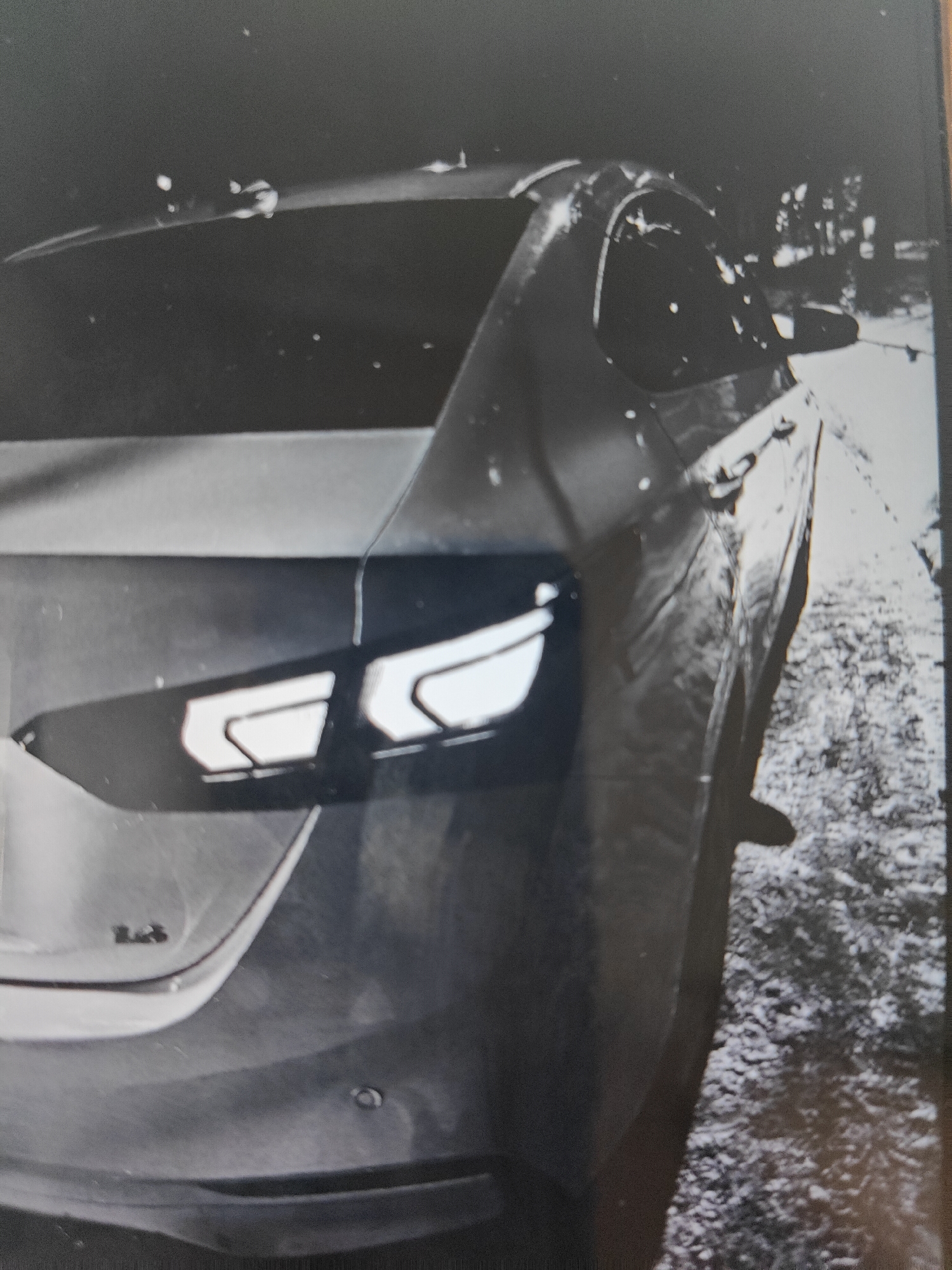 The image shows the rear driver's side of a dark-colored sedan, likely a Chevrolet. The vehicle is parked outdoors at night, with evidence of patchy snow or ice on the ground. The car's taillight assembly is visible and appears to have a distinctive 'C' shaped LED pattern. The body of the car shows some surface imperfections, possibly minor damage or dirt. The background is dark and somewhat blurry, suggesting a low-light environment with limited visibility. No people or specific landmarks are identifiable in the image that would indicate a precise location within Pushkino, Russia.