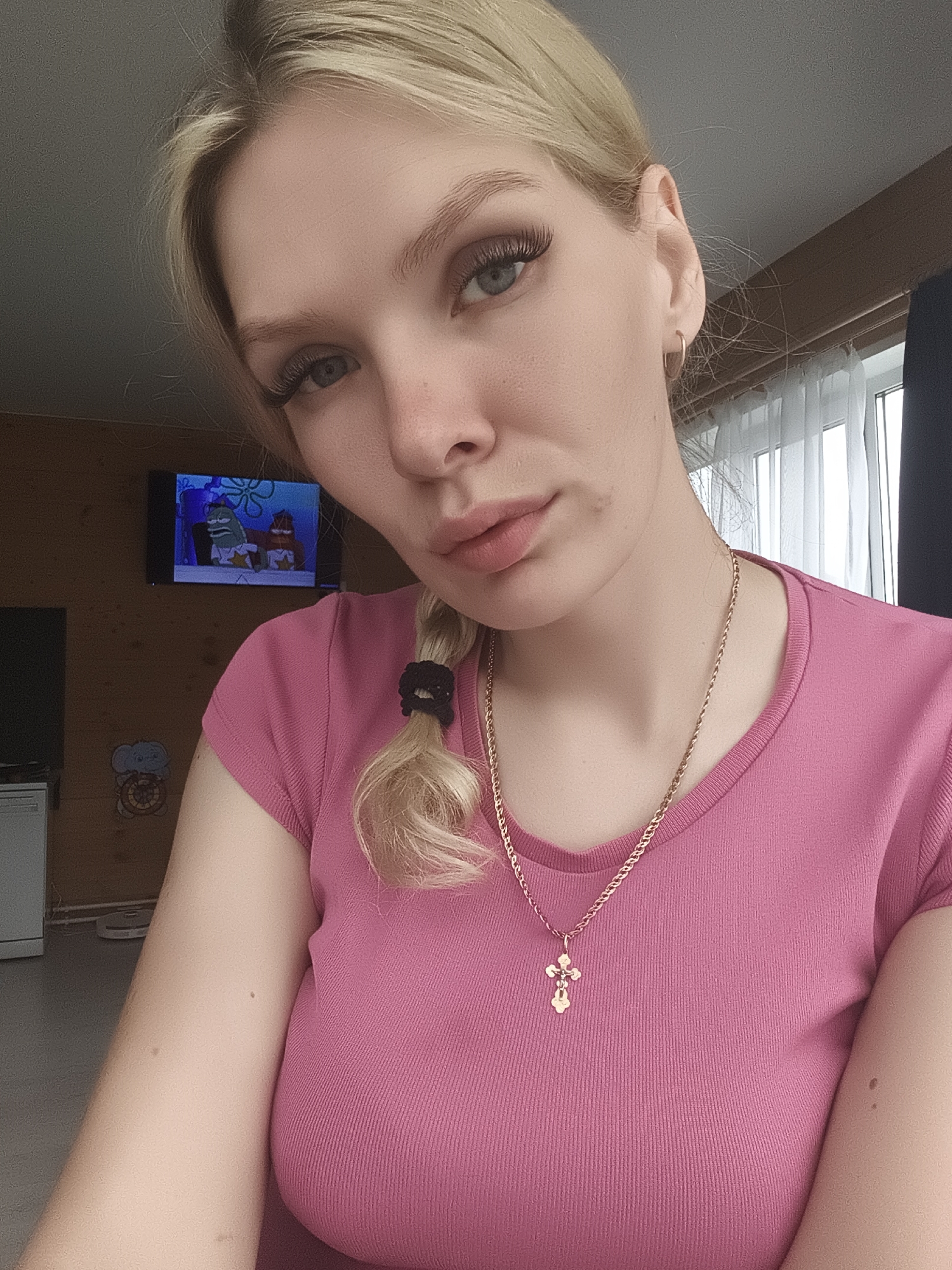 The image is a close-up, indoor selfie of a young Caucasian woman with blonde hair braided to her left. She is wearing a pink t-shirt and a gold cross necklace. Her eyes are blue, and she has long eyelashes. Her lips are slightly parted, and she has a beauty mark on her cheek. In the background, a television screen shows a cartoon, and a window with white curtains is visible. The overall setting appears to be a home environment. The location is identified as Русский Вожой, Russia.