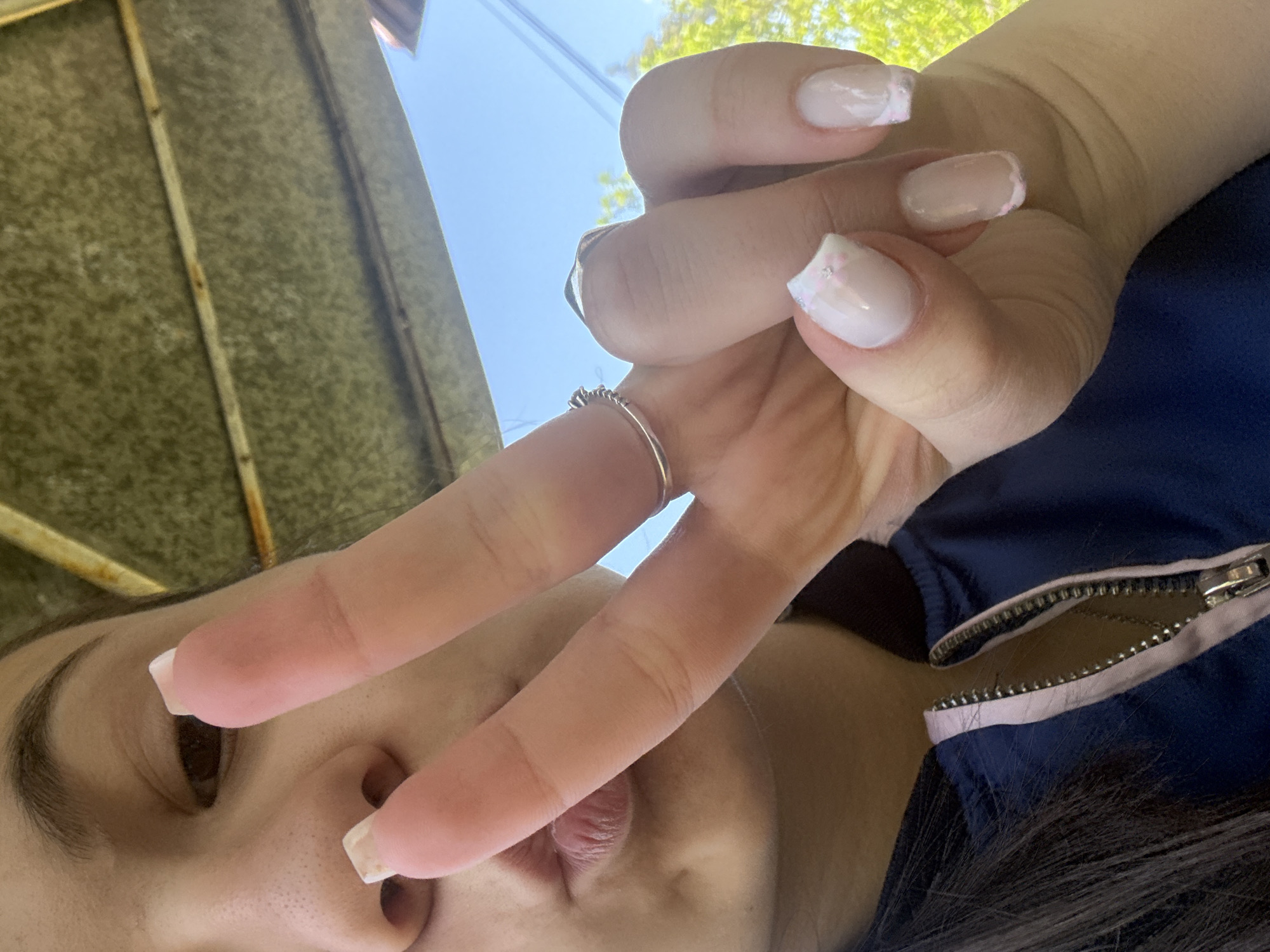 The image is a selfie taken from a low angle, focusing on a person's face and hand making a peace sign. The person has dark hair and light skin. They are wearing a silver ring on their middle finger. Their nails are manicured with a light pink base and white tips with iridescent glitter accents. The person is wearing a blue jacket with a zipper. The background includes a corrugated metal or textured surface, possibly a roof or awning, and a patch of blue sky with some green foliage visible, suggesting an outdoor setting. The location is not definitively discernible as Radomir, Bulgaria from the visual information provided.