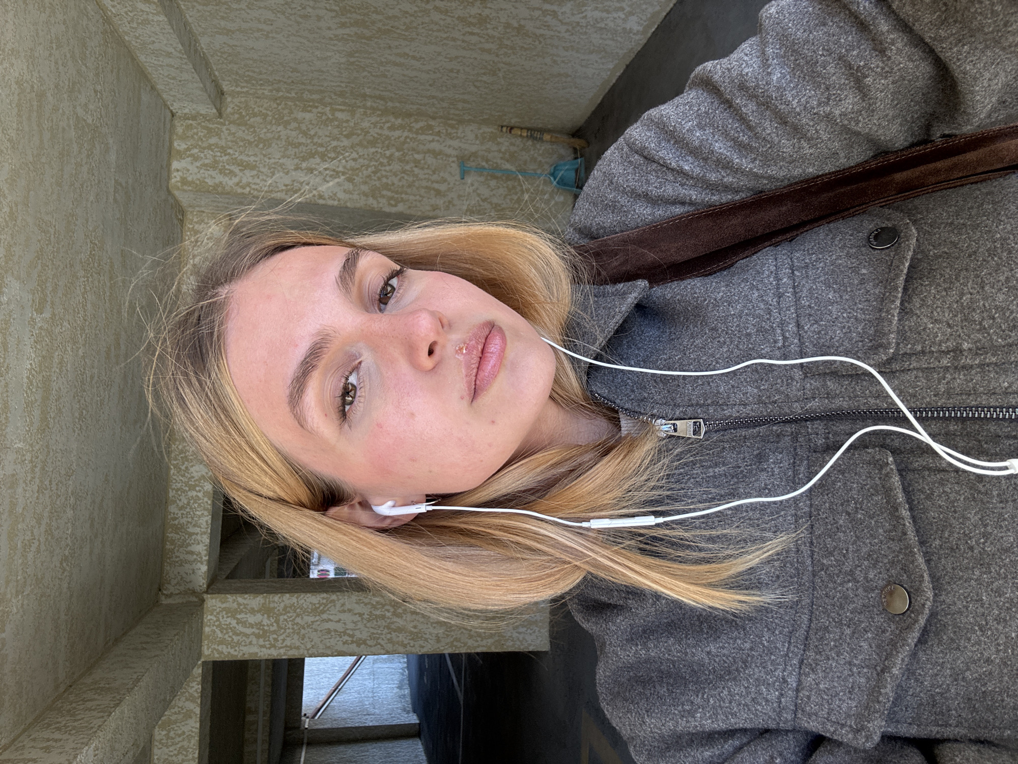A young woman with blonde hair wears a gray coat and white earphones. She's wearing a dark brown shoulder strap. She is standing in a concrete alcove looking at the camera.