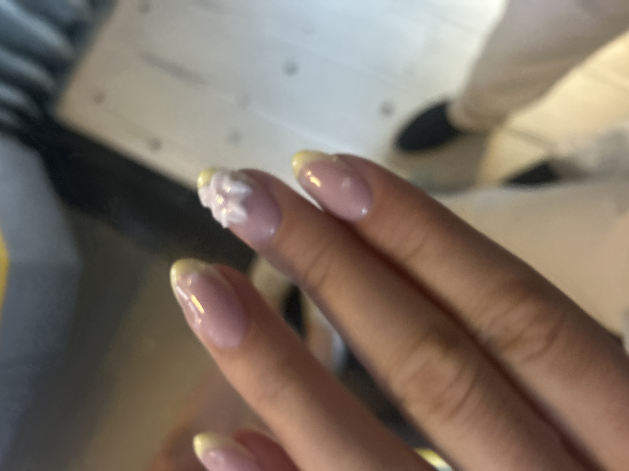 The image shows a close-up view of a person's hand with manicured nails. The nails are French manicured with a sheer pink base and a pale yellow tip. One nail has a small, three-dimensional white embellishment that resembles a flower. The background is blurred, but appears to be an indoor setting with a floor that has a grid-like pattern. A dark shoe and light-colored pants are visible in the lower right portion of the frame, suggesting the presence of another person or the photographer's own attire. The lighting is soft, and the overall impression is one of personal grooming and a casual setting. The location is not identifiable from the image.