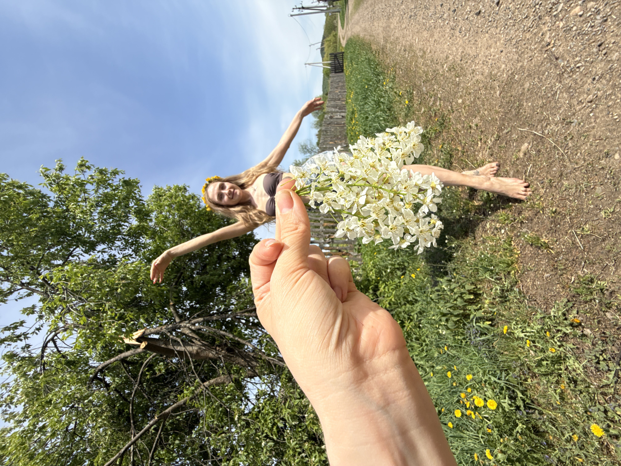 The image shows a woman with long blonde hair wearing a brown bra and a yellow floral crown, barefoot and reclining near a dirt road in Nizhnyaya Chermoda, Russia. She has her arms outstretched, and a hand holding a bouquet of white flowers is in the foreground. A tree is on the left, and a dirt road runs into the background. There is a fence on the right side of the road, and power lines are visible in the background. Dandelions grow in the green grass. The sky is partially cloudy and blue.