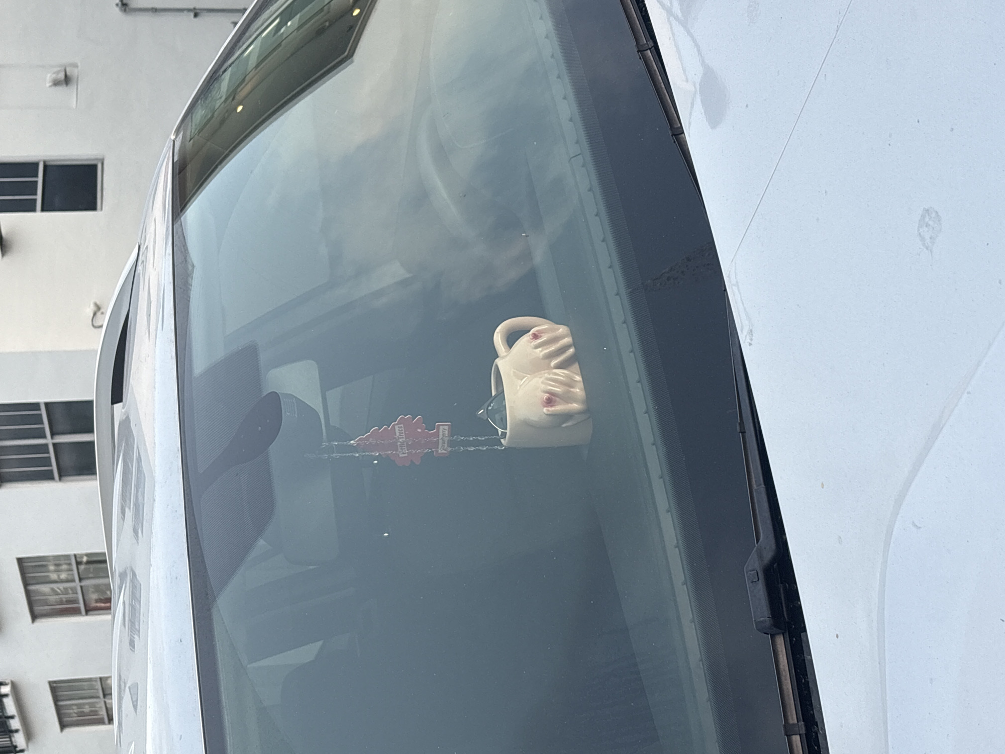 An angled, close-up shot captures the front windshield and part of the white body of a vehicle, likely a car or van, parked outdoors in what appears to be an urban or suburban setting in Miami Beach, USA. The daylight and clear reflections on the windshield suggest a bright, sunny day.
Through the windshield, two distinct objects are visible within the vehicle's interior. On the dashboard or hanging near the rearview mirror, there is a novelty light-colored mug or cup. This mug is uniquely shaped like a human torso, with molded hands cupping the breast area, featuring prominent pink nipples. A pair of dark sunglasses is casually placed inside this mug. To the left of the mug, a red, tree-shaped "Little Trees" brand air freshener hangs. The text "Little Trees", "Air Freshener", and the scent name "Strawberry" are clearly legible on it.
In the background, a plain white, multi-story building with several rectangular windows is visible, reflecting a common architectural style. Parts of the vehicle's white hood or roof show minor smudges or dust. A black windshield wiper is also visible on the right side of the frame. The scene is static, showing no explicit activity, but rather a snapshot of a parked vehicle with its personal interior decorations.
