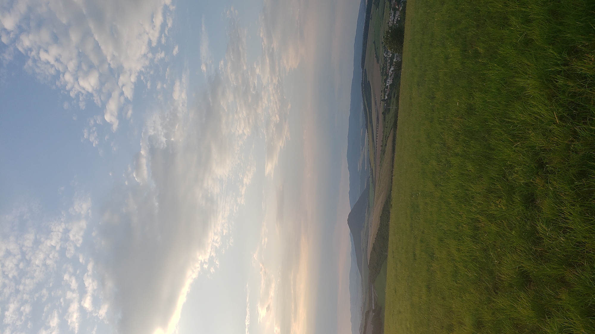 This is a landscape photograph taken in the city of Prešov, Slovakia. The foreground is dominated by a lush, green grassy field, illuminated by the soft light of a setting or rising sun. In the mid-ground, rolling hills and fields stretch out, with a distinct conical hill rising prominently in the center. Beyond these, a hazy mountain range forms the distant horizon. The sky above is a mix of pale blue and scattered white clouds, with a bright, diffused light source suggesting a time near sunrise or sunset. A small village or town is visible in the distance, nestled among the hills. The overall impression is one of a serene and expansive natural landscape with human settlement in the distance.