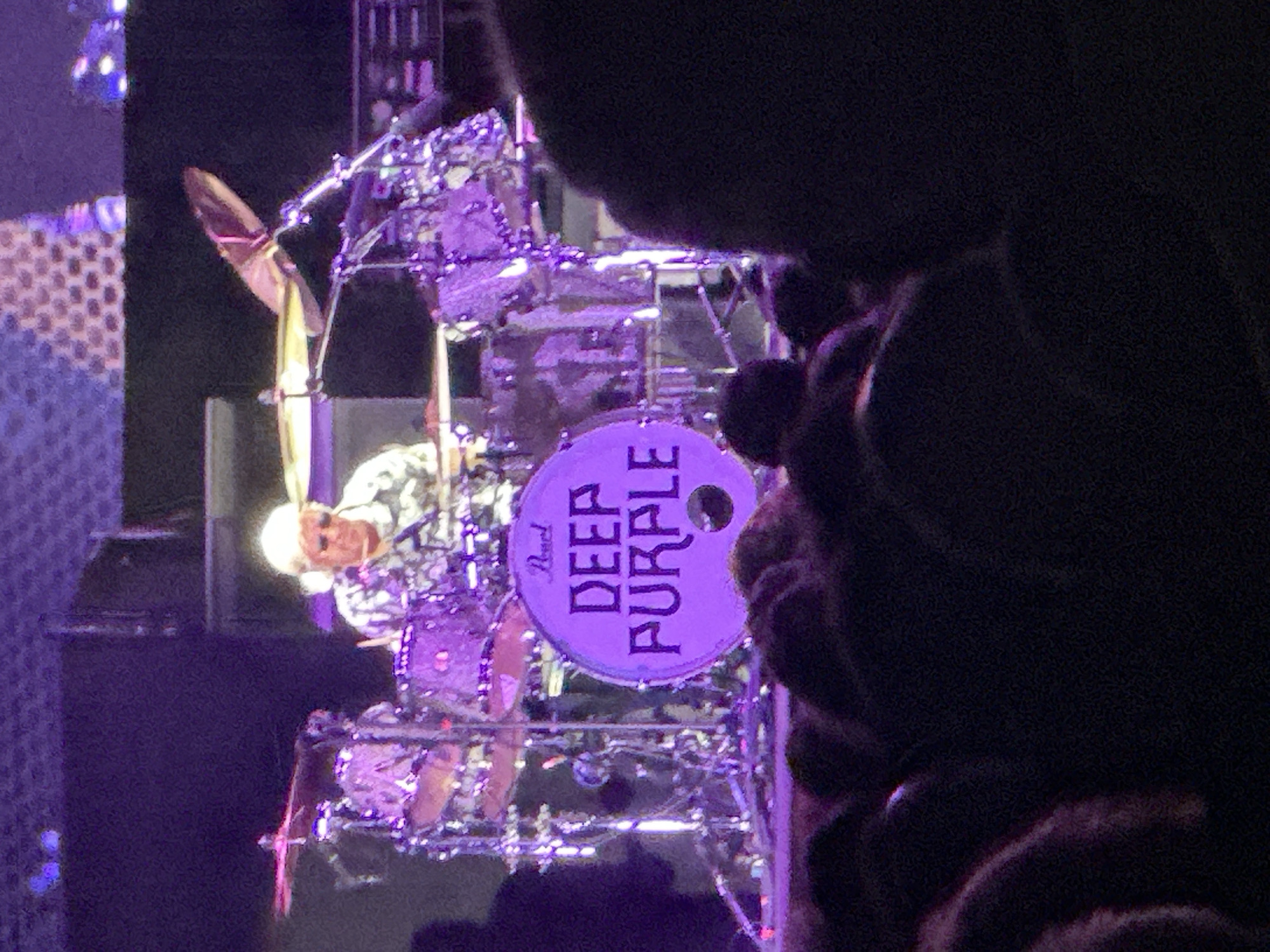 The image shows a drummer playing a drum kit on a stage. The drummer is wearing a white and black patterned shirt and sunglasses. The drum kit is illuminated by purple stage lights. The kick drum has the words "DEEP PURPLE" written on it in purple letters with a white outline. The background is dark, with some stage equipment visible. The audience is blurred in the foreground and appears to be wearing dark clothing. This image was taken at the concert hall in Jerez De La Frontera, Spain.
