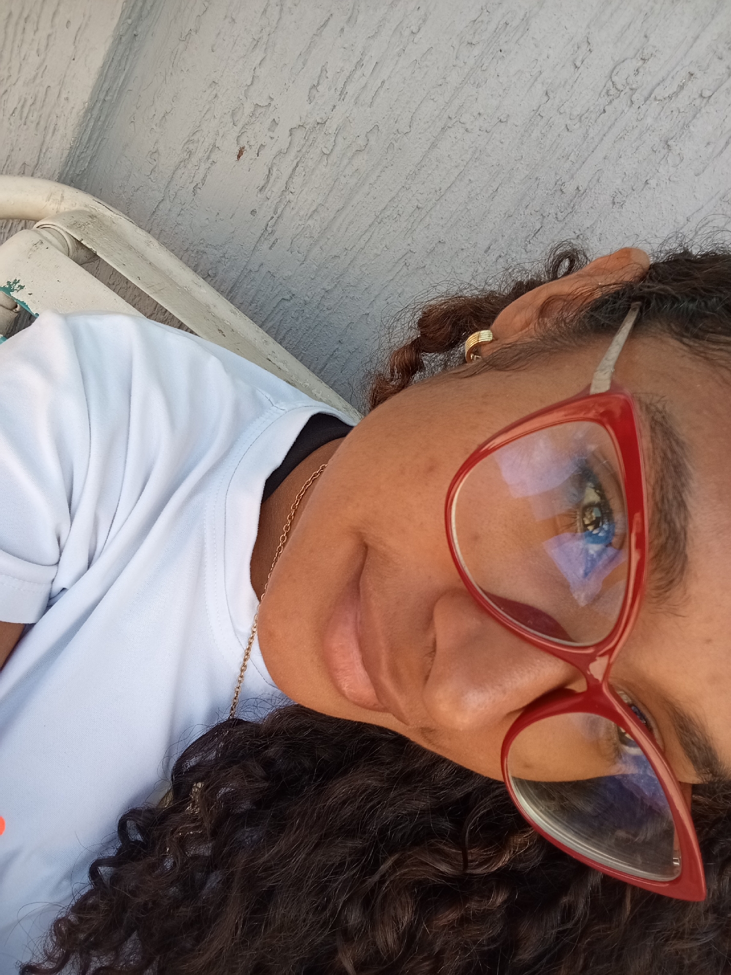 A medium close-up portrait captures a woman positioned at an angle, with her head tilted to the right. She has a medium-dark complexion and dark, curly hair. Her facial expression shows a subtle, gentle smile, conveying a calm demeanor. She is wearing stylish red-framed glasses, and hints of blue and purple are reflected in the lenses, possibly from the sky or surroundings. A small gold hoop earring is visible on her right ear, and a thin gold chain necklace rests against her white t-shirt.
She appears to be seated outdoors, with a fragment of a light-colored, possibly white or off-white, chair or bench visible in the lower left of the frame. The background features a light-colored wall with a distinct rough, textured surface, likely stucco, showing some areas of worn or peeling paint. The bright lighting suggests it is daytime. The scene evokes a casual outdoor setting in Juan Griego, Venezuela. No text is visible in the image.