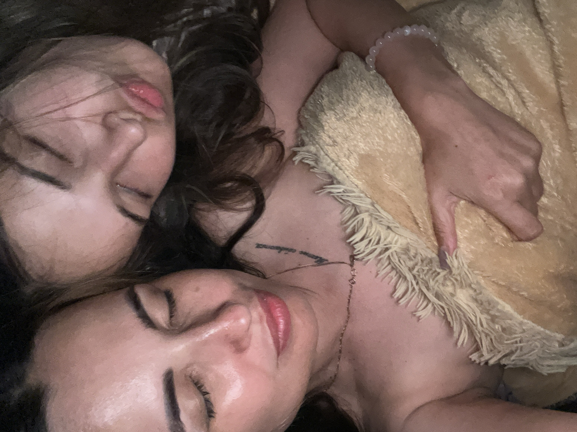 The image shows a close-up, overhead shot of two people lying down, their faces close together, with their eyes closed. They appear to be sleeping or resting. One person's face is in the upper left portion of the frame, with dark hair around their head. Their lips are slightly parted and appear to have lipstick on. The other person's face is in the lower part of the frame, also with their eyes closed. They have makeup on, including eyeliner and lipstick. A tan-colored blanket with fringe is visible in the background, covering part of the scene and one person's arm. The arm has a bracelet made of light-colored beads. A delicate necklace is also visible around the neck of the person in the lower part of the frame. There is faint text visible as a tattoo on the neck of the person in the lower part of the frame. The lighting is dim, suggesting it could be nighttime or a dimly lit room. The overall emotion conveyed is one of peace and intimacy.