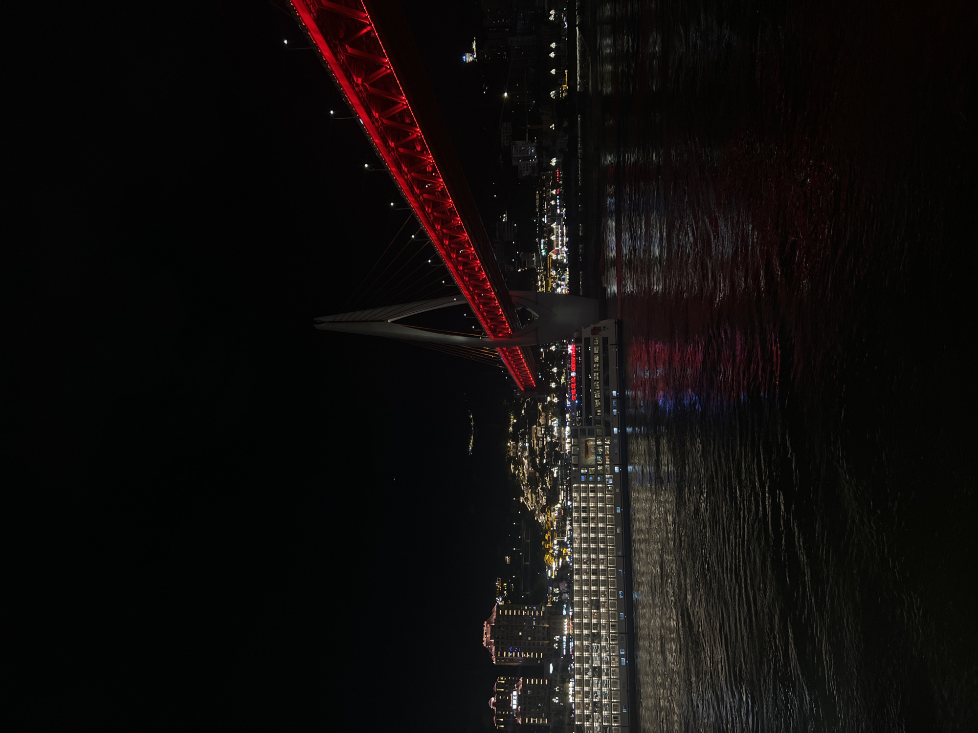 An eye-level, long shot captures a nighttime cityscape in Yuzhong District, China, dominated by the illuminated Hongyadong Bridge and a large riverfront. The bridge, a prominent red suspension structure, spans the width of the image from right to left, with its support cables and roadway brightly lit in red. Below the bridge, the river reflects the vibrant red lights, creating a shimmering effect on the dark water.
Along the riverbank, a multi-story cruise ship is docked, its many windows illuminated, casting a warm glow onto the water. Behind the ship, the Hongyadong area is visible, a hillside densely packed with traditional buildings and illuminated windows, appearing as a glittering cascade of lights. Several taller modern buildings are also visible in the background, their lights adding to the urban glow. The sky is a deep black, with only a few faint stars visible. No people are discernible in the image.