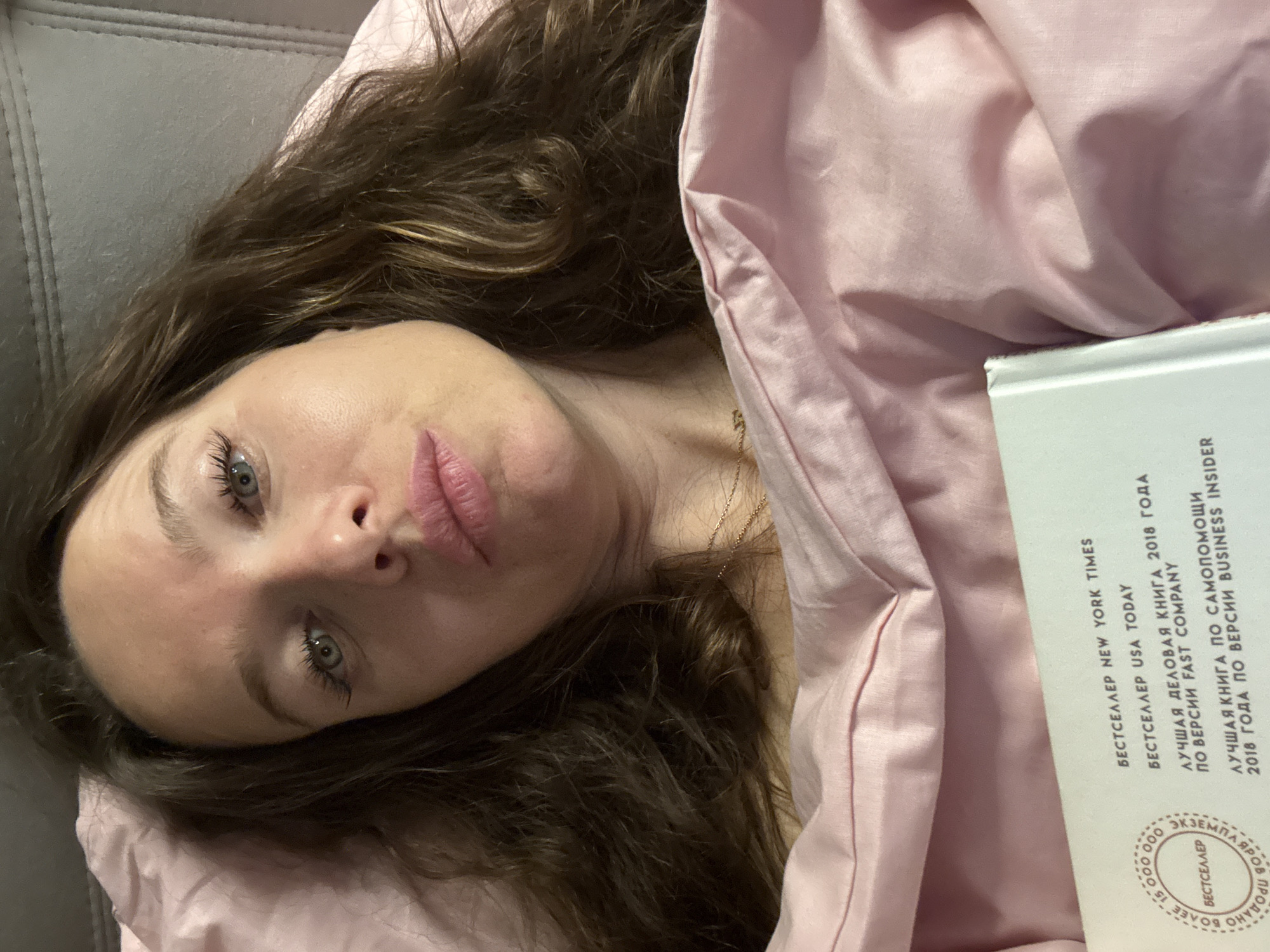 A woman is depicted lying on her back, looking towards the viewer, in an indoor setting in Khimki, Russia. She has long, wavy dark brown hair, light blue-green eyes, and her lips are slightly pursed. A delicate gold chain necklace is visible around her neck. She is partially covered by a soft, light pink blanket or duvet, with a grey upholstered surface visible behind her head, possibly a couch or headboard.
A white or light grey book is placed beside her, tucked into the blanket. The visible cover of the book features text in both English and Russian, indicating it is a bestseller. The English text reads: "NEW YORK TIMES BESTSELLER" and "USA TODAY BESTSELLER". The Russian text translates to: "BESTSELLER NEW YORK TIMES", "BESTSELLER USA TODAY", "BEST BUSINESS BOOK OF 2018 ACCORDING TO FAST COMPANY", and "BEST SELF-HELP BOOK OF 2018 ACCORDING TO BUSINESS INSIDER". A circular stamp also visible on the cover indicates "BESTSELLER" and "MORE THAN 150,000 COPIES SOLD" (БОЛЕЕ 150 000 ЭКЗЕМПЛЯРОВ ПРОДАНО). The woman appears relaxed, perhaps resting or pausing from reading. The lighting suggests an indoor environment.