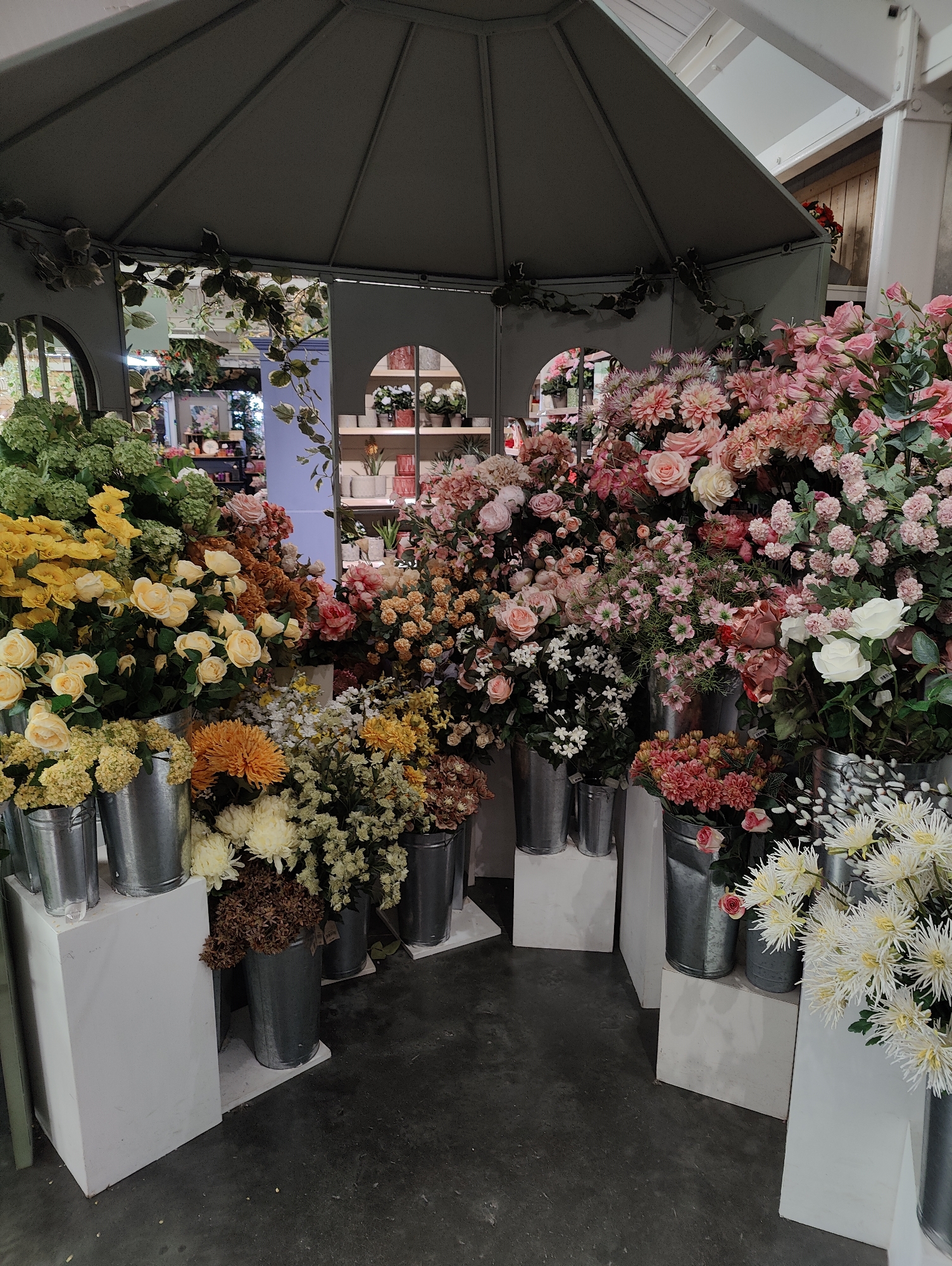 The image depicts an indoor display of artificial flowers, arranged under a gray, gazebo-like structure. The flower selection is abundant and diverse, featuring a range of colors including yellow, orange, white, and various shades of pink and red. The flowers are arranged in metallic buckets which are placed on white rectangular stands. The flooring is a dark gray, and the backdrop reveals glimpses into the surrounding interior, including shelves with additional decorative items. The scene is located in Colchester, United Kingdom.