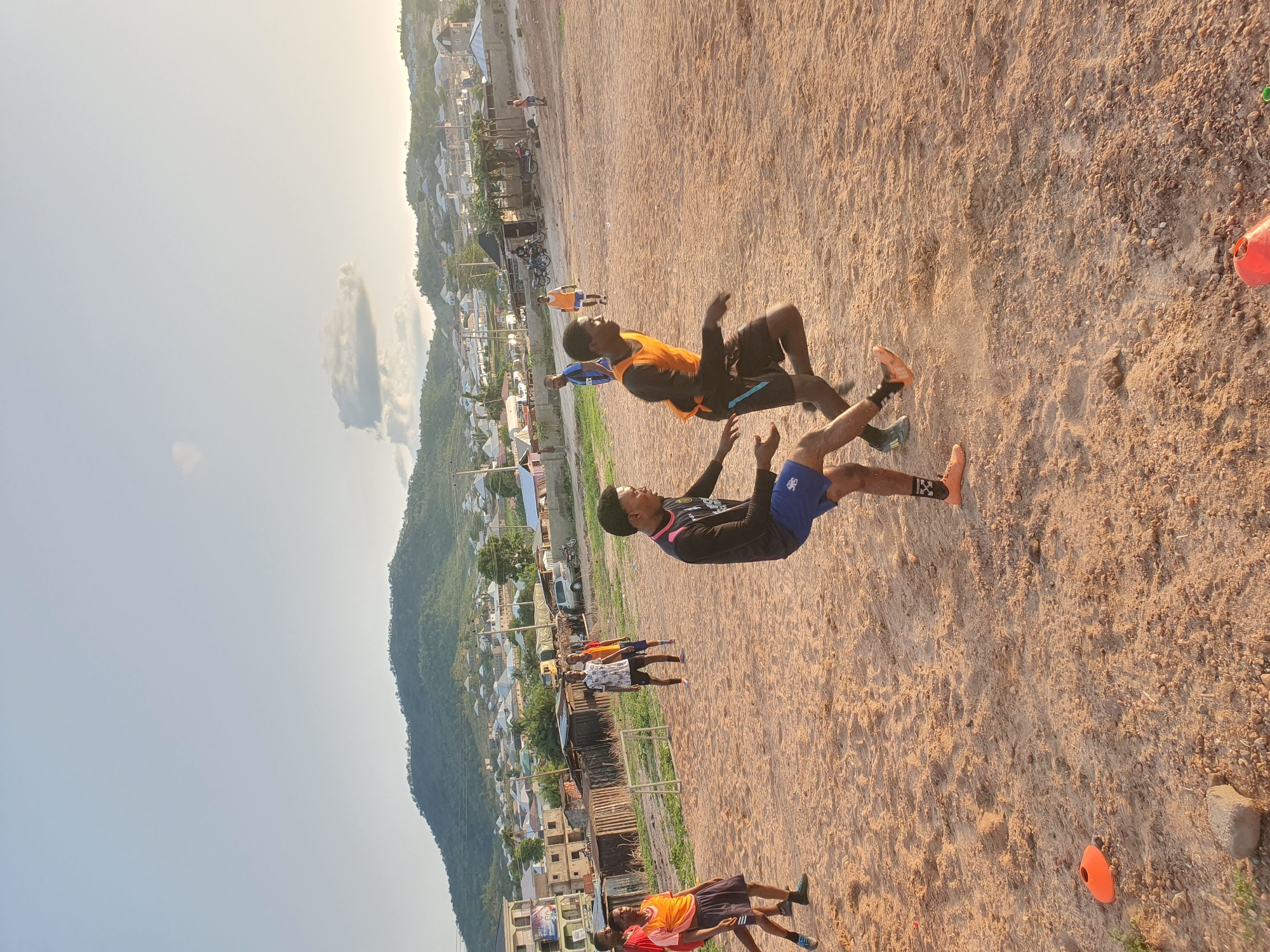 The image shows a sandy field in Lokoja, Nigeria, with a backdrop of a hillside community. Two men are in mid-air, appearing to practice or demonstrate flips. One man wears an orange athletic vest over a black long-sleeved shirt and black shorts with orange shoes. The other wears a black long-sleeved shirt with blue shorts and peach shoes. Other people stand on the field, some wearing similar athletic attire. Orange cones are also visible. The time of day seems to be late afternoon or early evening due to the long shadows.