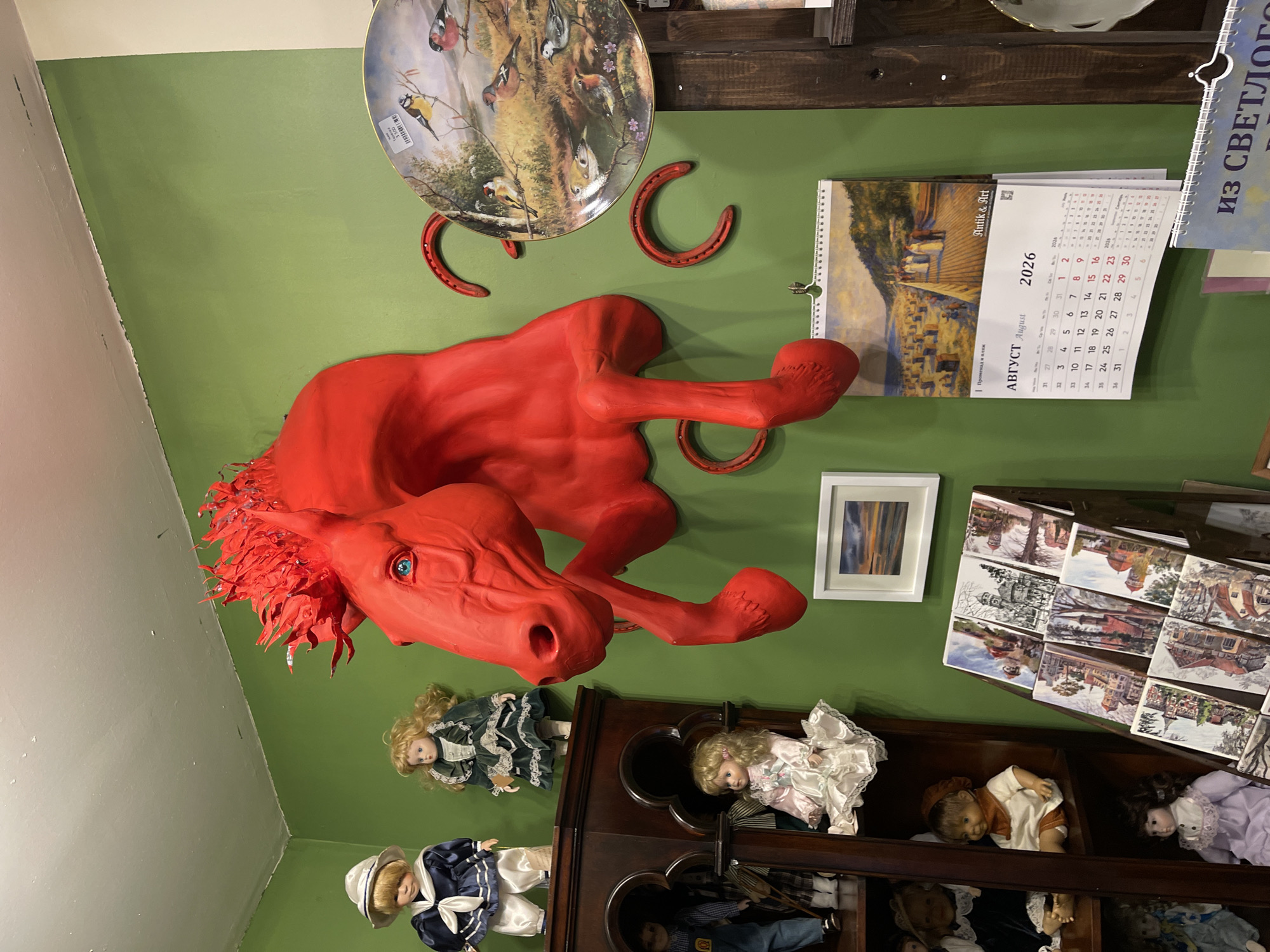 The image shows a room with green walls, likely a shop or a private collection, filled with various decorative items. Prominently displayed on the wall is a large, bright red sculpture of a horse's head and upper body. The horse's eyes are a striking blue. To the right of the horse sculpture, a decorative plate featuring birds in a natural setting is hanging on the wall. Below the plate, two red horseshoe decorations are affixed to the wall. A calendar for August 2026 with a seaside scene is also hanging on the wall.
On shelves to the left, several porcelain dolls are displayed. One doll in a sailor outfit is on the top shelf. Next to it, another doll in a dark green dress is also visible. Below them, more dolls are arranged on lower shelves, including a doll in a white dress and another in a purple dress. A display rack filled with what appear to be postcards or small prints of buildings and landscapes is placed in front of the shelves. A small framed artwork depicting a sunset over water hangs on the wall between the horse sculpture and the doll display. A wooden display unit with dark wood is to the right of the shelves.
The text on the calendar indicates "АВГУСТ August 2026" and shows a beach scene with people and beach huts, which is evocative of coastal resort towns. The phrase "из СВЕТЛОГО" ("from Svetly") is partially visible on a calendar at the bottom right, suggesting a connection to or origin from Svetly, Russia. Given the presence of beach-themed imagery on the calendar and the mention of Svetly (which is a city in Kaliningrad Oblast, Russia, located on the Baltic Sea), it is plausible that this scene is located in or related to the city of Svetlogorsk, Russia, which is also a well-known Baltic Sea resort city in the same region and is often associated with similar artistic and decorative elements.