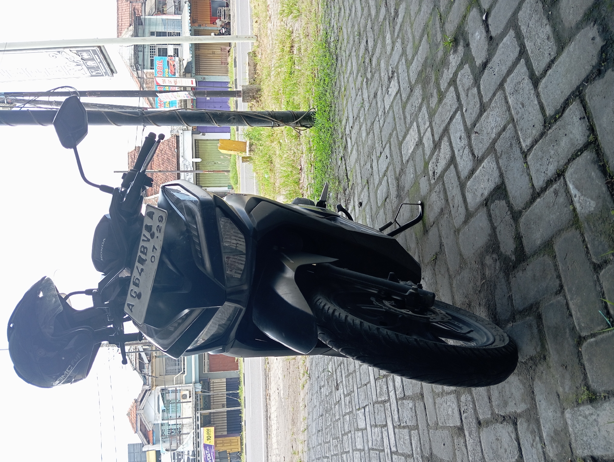 The image depicts a black Honda Vario scooter parked on a grey paver brick surface, laid in a herringbone pattern, exhibiting patches of dirt and sparse green vegetation. A black SMC Fuji helmet rests on the scooter's handlebars, positioned above the visible front license plate, "AG 544BV 07.29". To the right of the scooter, a dark utility pole, wrapped with cables, stands within a strip of green grass and dirt. Beyond this strip lies a two-lane asphalt road. Across the road, a row of commercial buildings with mostly closed, roll-down metal shutters in various colors (green, purple, brown, yellow) forms the background. Advertisements are visible on these buildings, including a "VOLT 10.000" banner on the far left and a "Jual Beli Mobil" billboard featuring a red car on the mid-right. Two individuals are seated in front of a shop on the far right side of the road. The sky above is bright and overcast. The scene is located in the city of Pelem, Indonesia.