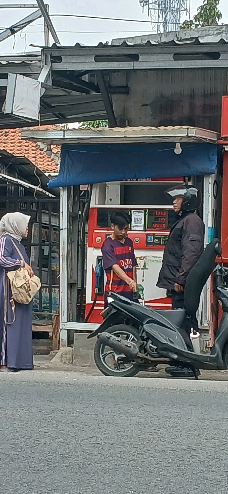 This is a medium-shot, full-frame photograph taken outdoors in Cirebon, Indonesia, depicting a scene at a gas station. The primary focus is on three individuals and a motorcycle. On the left, a person wearing a hijab and a blue abaya stands facing right, holding a light-colored bag over their right shoulder. In the center, a young man in a purple and red striped t-shirt is at a gas pump, holding a green gas nozzle. To the right, a person wearing a black jacket and helmet is standing and facing the young man. A black motorcycle is parked in the foreground, angled towards the right of the frame, with its kickstand down. Behind the people is a gas station structure with red and white accents, a blue tarp overhead, and visible gas price displays. The background shows a building with a tiled roof and structural elements, possibly part of a shop or shelter. The ground is asphalt. The overall lighting suggests an overcast day.