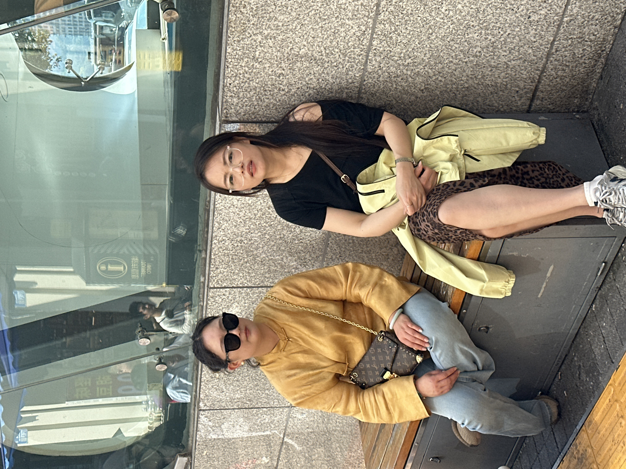 Two women are seated next to a glass window in an urban area. One woman wears glasses, a black top, a leopard print skirt, and a light yellow jacket. The other woman wears sunglasses, a gold-colored top, and jeans. They both have bags. The scene appears to be in Yuzhong District, China, based on a Chinese information sign visible in the glass.