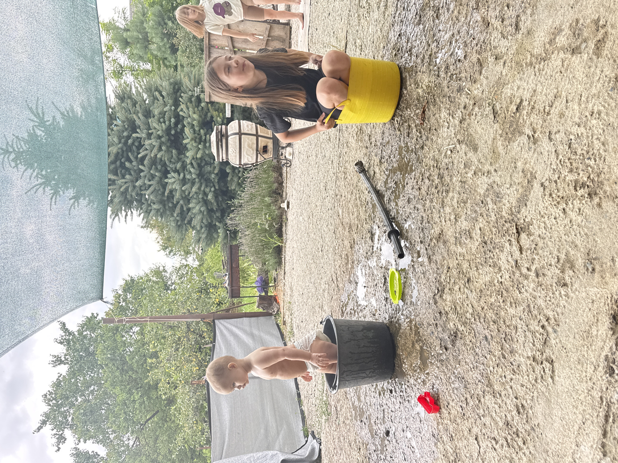 In a backyard setting in Zassovskaya, Russia, two children are engaged in a water play activity. A young girl, with long brown hair and wearing a black t-shirt and shorts, is sitting in a bright yellow bucket, holding onto its handles. Her facial expression suggests she is enjoying herself. In the foreground, a baby, wearing only a diaper, is sitting inside a large black bucket, with its legs dangling outside. The ground around the children is wet, indicating water has been spilled or splashed.
Scattered on the wet ground are a few plastic toys: a green colander-like object and a small red toy. A water hose nozzle is also visible. Behind the children, trees and greenery are abundant, suggesting an outdoor, possibly summer, day. A large tan-colored outdoor oven or grill is partially visible in the background, along with some wooden structures and a green shade canopy. The weather appears to be overcast, but the lighting suggests it is daytime. No visible text is present in the image.