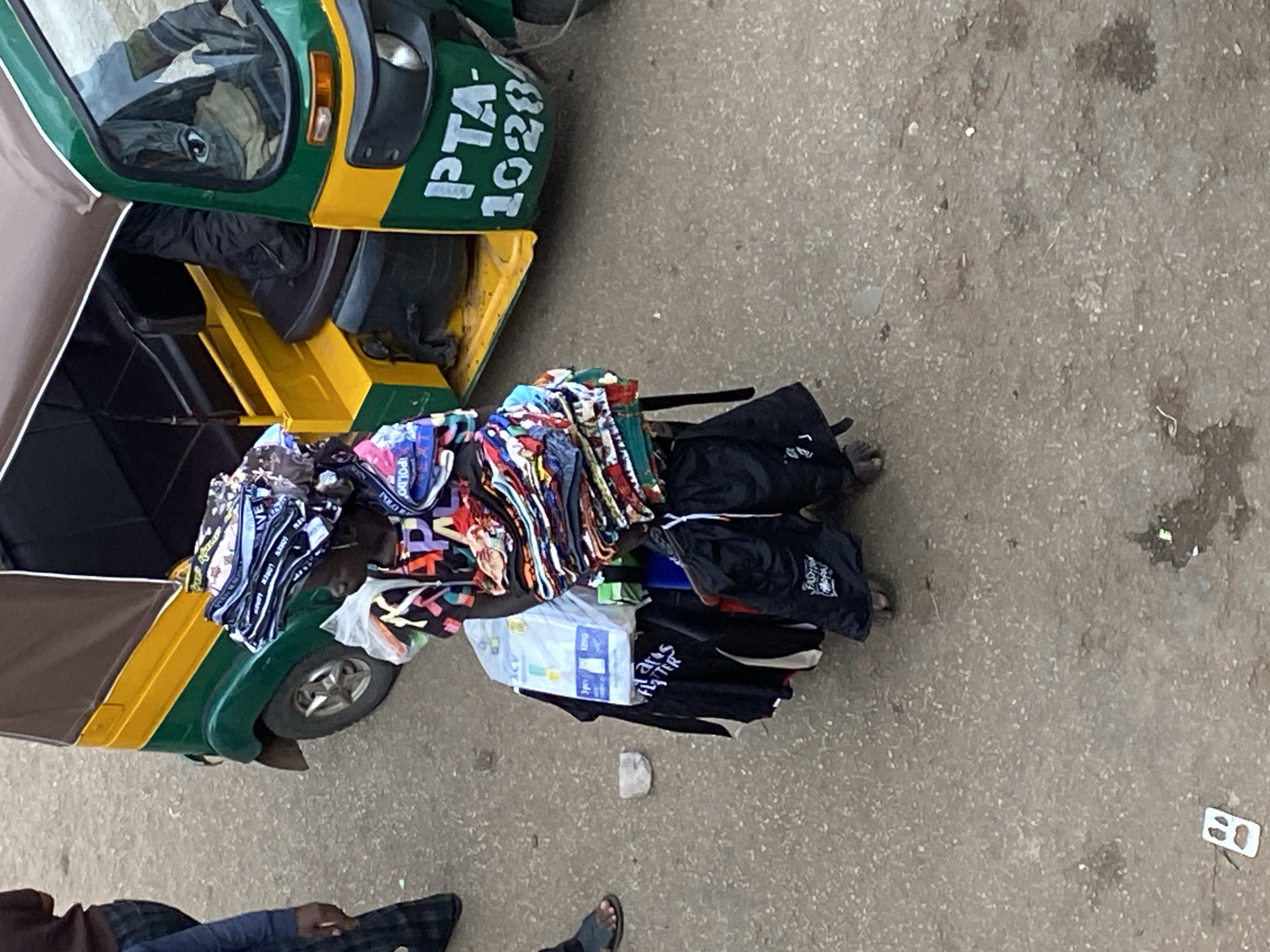 The image depicts a three-wheeled vehicle, possibly a Bajaj RE or similar model, parked on a concrete or asphalt surface. The vehicle is painted in green and yellow, with the registration number "PTA-1028" visible on the front. On the vehicle's roof and cargo rack, there is a pile of what appears to be clothing or textiles, including some branded items like "POLO." There is also a package of diapers or wipes.
A person is driving the vehicle. Another person stands near the vehicle, visible from the knees down. The setting is a road or street, potentially in Bukuru, Nigeria, based on the query.