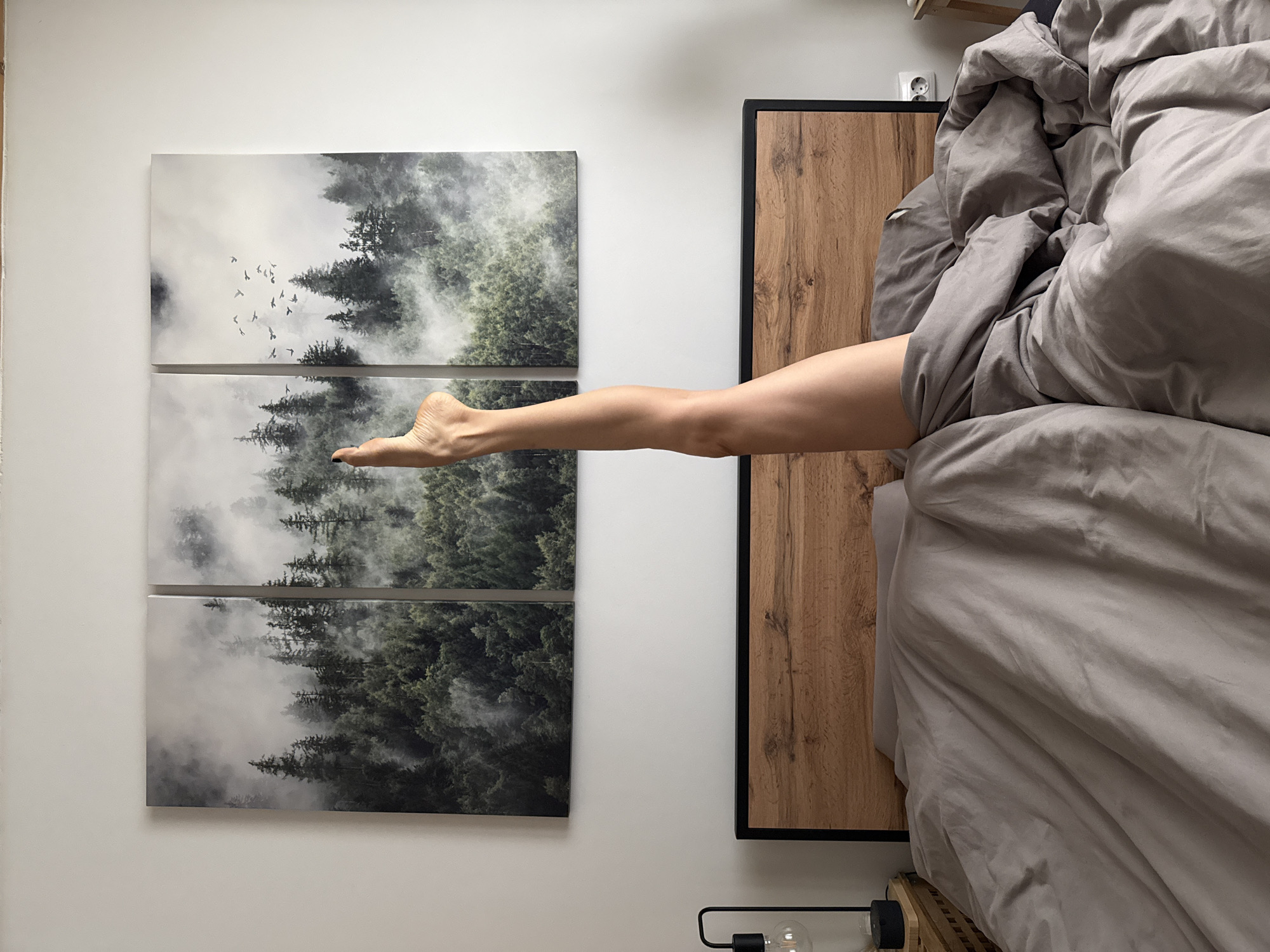 A woman's bare leg is propped up in the air from under a gray duvet on a bed. The bed has a wooden headboard and is positioned against a white wall. Above the headboard, there are three framed canvases of a misty forest. A black lamp with a clear bulb is on a nightstand to the left of the bed. The scene is indoors and appears to be a bedroom.