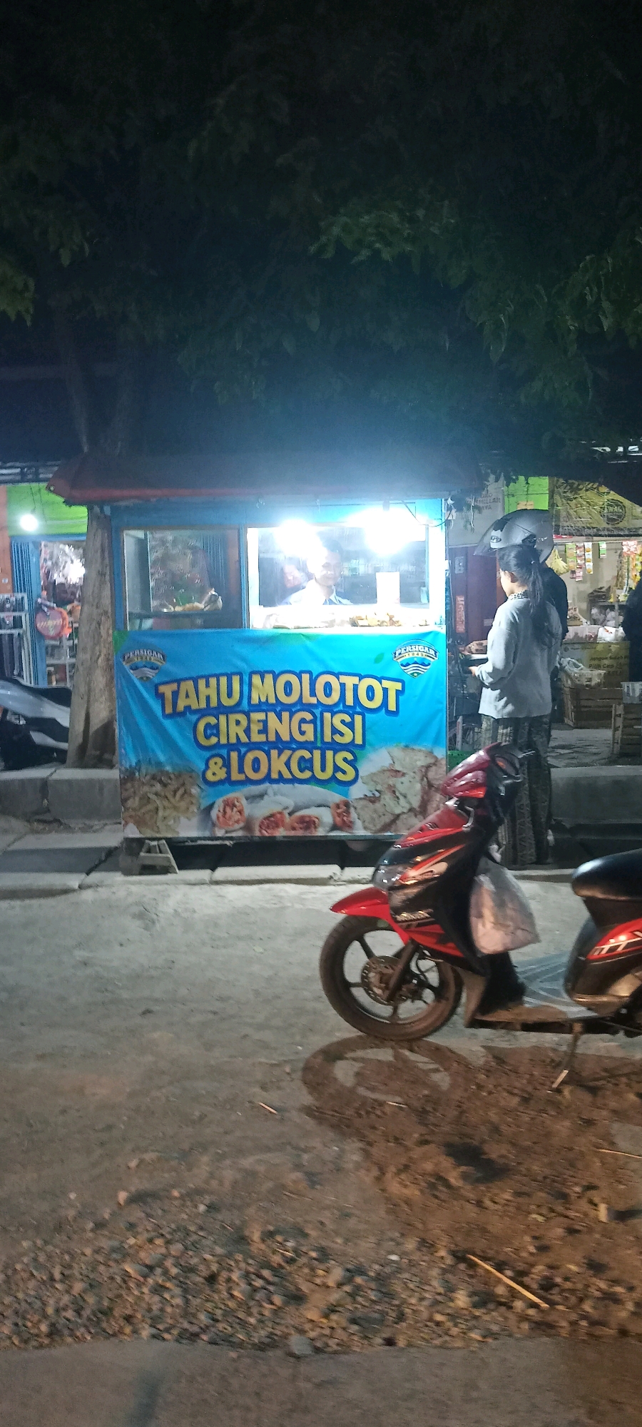 In Singogalih, Indonesia, a night scene unfolds with a food stall as the central focus. The stall, illuminated by bright lights, features a blue banner that reads "TAHU MOLOTOT CIRENG ISI & LOKCUS." Behind the counter, a man in a white shirt is visible, preparing food. A woman, wearing a gray long-sleeved shirt and patterned pants, stands at the counter, looking towards the man.
In the foreground, two motorcycles are parked. One is a red and black scooter, and part of a black scooter is visible to its right. The ground is a mix of asphalt and gravel. Trees with dark leaves hang over the top of the scene, creating shadows. The overall atmosphere suggests a bustling street food operation in the evening.