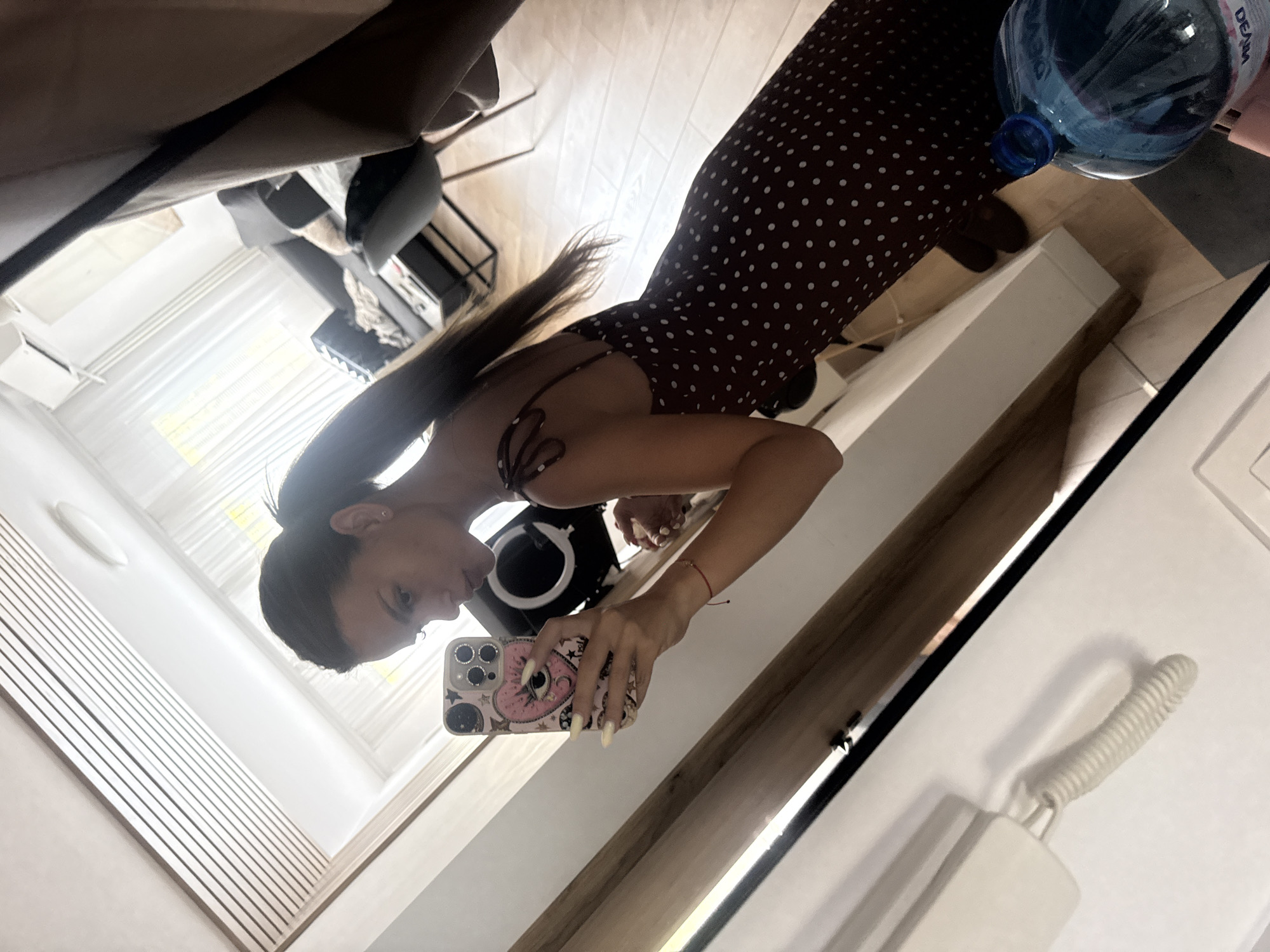 A young woman with dark hair pulled into a high ponytail is taking a mirror selfie in an indoor setting. She is wearing a dark red, polka-dotted dress with spaghetti straps. Her right arm is extended, holding a smartphone with a pink case decorated with a heart and eye design. She is leaning slightly forward, and her lips are pursed. In the background, a modern living room is visible, with a sofa, chairs, and a coffee table. There are sheer curtains covering a window, and the floor appears to be light-colored wood laminate. To the right of the woman, a large blue plastic bottle, possibly containing water, is visible. The overall impression is of a casual moment captured in a home environment, potentially in Plovdiv, Bulgaria, given the prompt's specific location context.