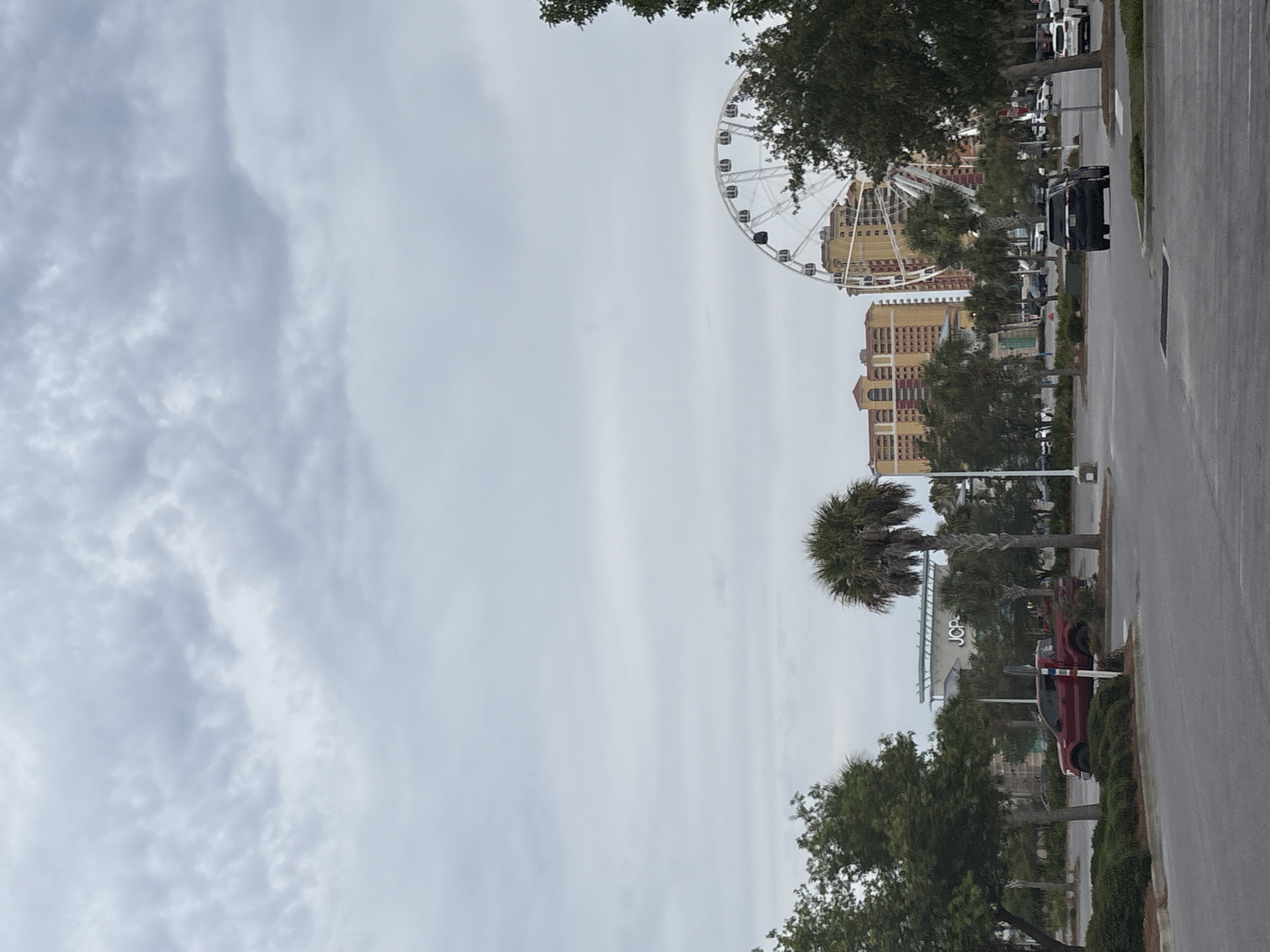 The image shows an outdoor view of a parking lot with several vehicles, including a red Ford F-150 pickup truck and a black SUV. In the background, there are buildings, palm trees, and a Ferris wheel. The sky is overcast with gray clouds. The JCPenney logo is visible on a building in the distance. This scene is likely in Panama City Beach, Florida, given the presence of palm trees, beach-style architecture, and the Ferris wheel, which is a common attraction in tourist destinations like Panama City Beach.