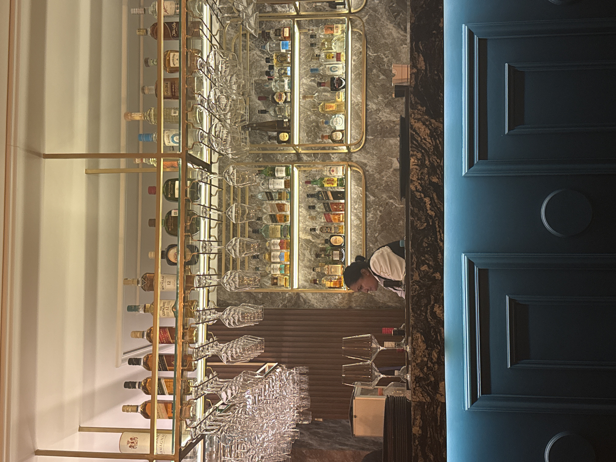 The image shows a well-stocked bar, potentially in Puebla, Mexico. The bar features multiple shelves displaying a variety of alcoholic beverages. Wine glasses hang upside down on metal racks. A person in a light-colored uniform is working behind the bar. A section of a blue wall with decorative moldings is visible on the right side of the image. The bar appears to be clean and organized.