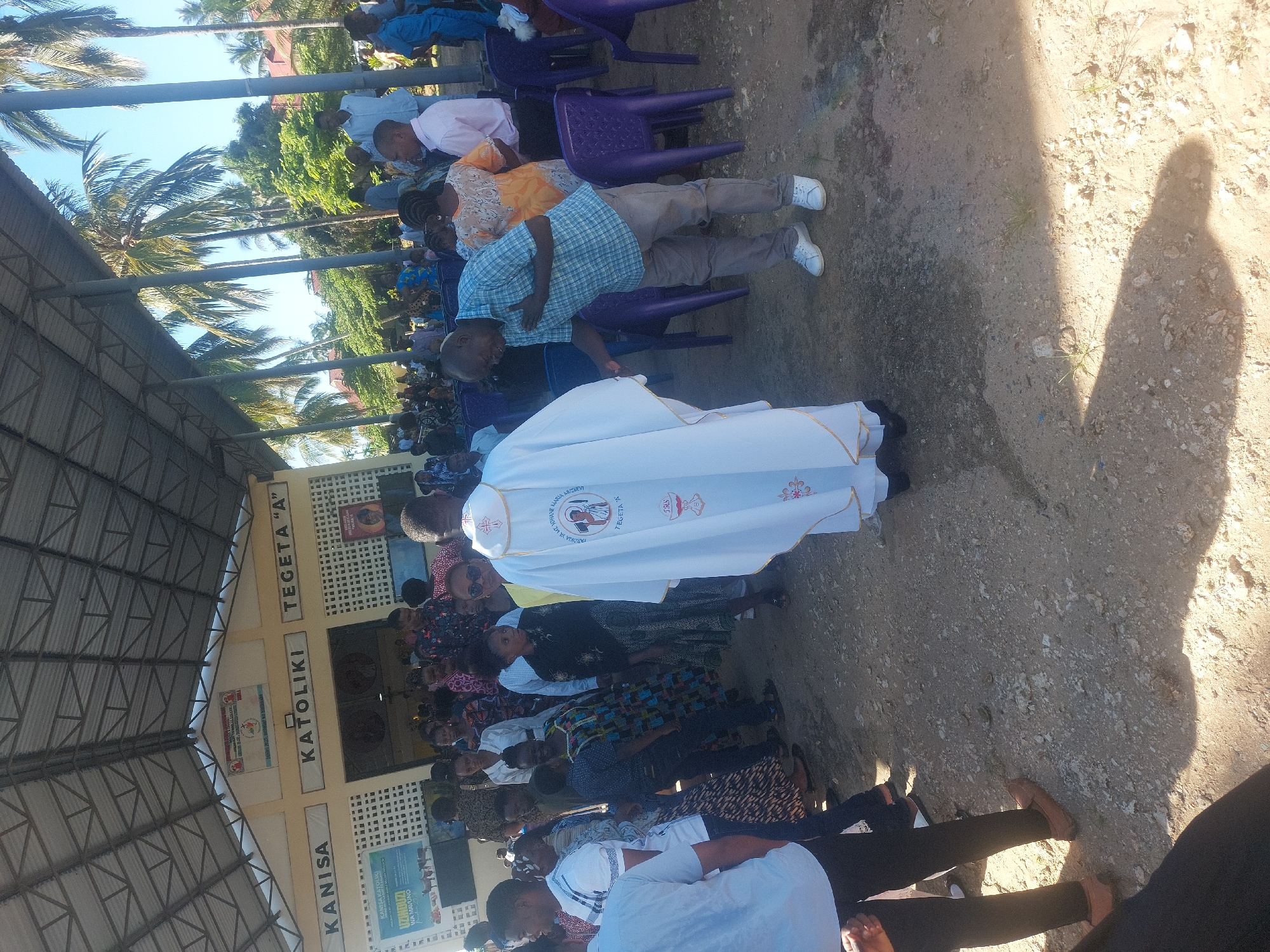 A medium-angle outdoor photograph captures a gathering of people, possibly a church service, at the Kanisa Katoliki Tegeta "A" (Catholic Church Tegeta "A") in Dar es Salaam, Tanzania. The image is oriented vertically.
People: The image contains many people. A man wearing a white robe with gold trim walks in the middle. Others, mostly in casual clothing, are seated in rows of purple plastic chairs. Some are standing.
Subjects: A church service or religious event is occurring.
Objects: The objects include the church building, identifiable by its signage. Rows of purple plastic chairs line the area. The man in white is the only one wearing religious garments.
Actions: The people are gathered together. The man in white is walking, likely leading or participating in the service. Others are standing or seated.
Interactions: The priest and others are interacting as part of the religious ceremony.
Environment: The gathering is taking place outdoors in an area next to a church building. Palm trees are visible in the background. The area appears to be a dirt or gravel ground.
Location: The location is Dar es Salaam, Tanzania.
---