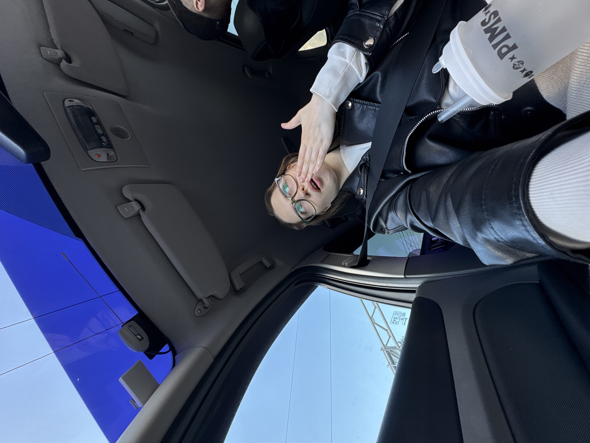 A medium shot captures a young woman with brown hair and glasses sitting in the passenger seat of a car, holding a white cup with a straw. She is wearing a black leather jacket and a white sweater. Her right hand is raised to her mouth, and her eyes are looking upwards. The car has a sunroof with blue tint, and the interior appears to be gray. Outside the car window, a bright blue sky is visible, with some overhead structures that could be part of a bridge or highway. The overall lighting suggests it is daytime. The scene is set in Saint Petersburg, Russia.