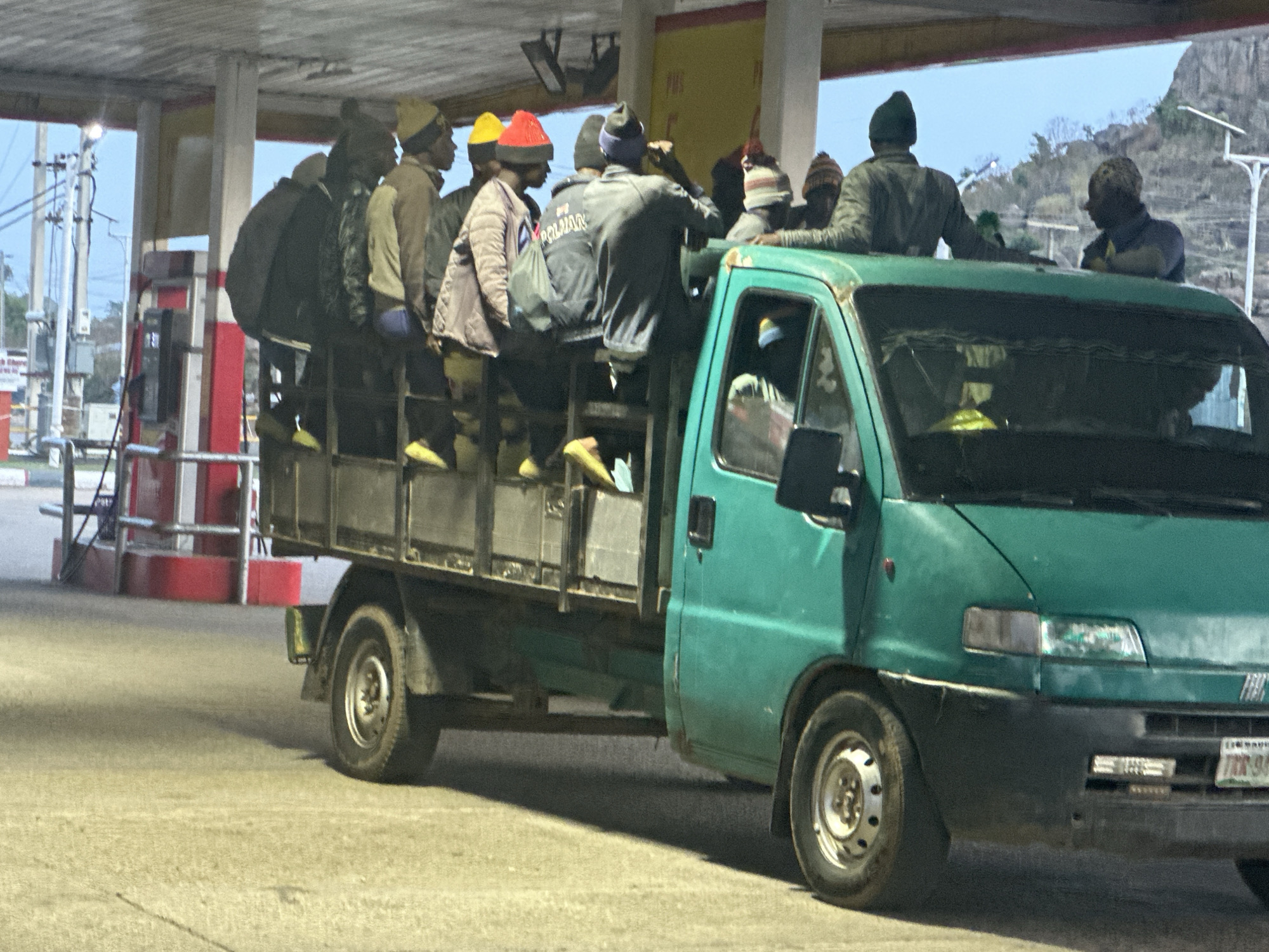 A green utility vehicle, resembling a Fiat Ducato-style van-based truck, is seen at what appears to be a gas station in Gura Top, Nigeria. Approximately 10-15 men are riding in and on the vehicle. Most are standing packed together in the open cargo bed at the rear, some wearing colorful knit hats (yellow, red, blue) and casual clothing. At least two men are also riding on the roof of the truck's cab.
The vehicle has a partially visible license plate on the front bumper that reads "TRB 94". In the background, to the left, are the red and white structures of a gas pump area, along with electrical poles and wires. The distant background shows a hilly or mountainous landscape under a light, clear sky. The lighting suggests it is either late afternoon, early evening, or early morning, with some areas shadowed. On the back of one man's jacket, the word "POLMAN" is visible. The scene depicts a common form of transportation in some regions, with the vehicle heavily loaded with passengers.