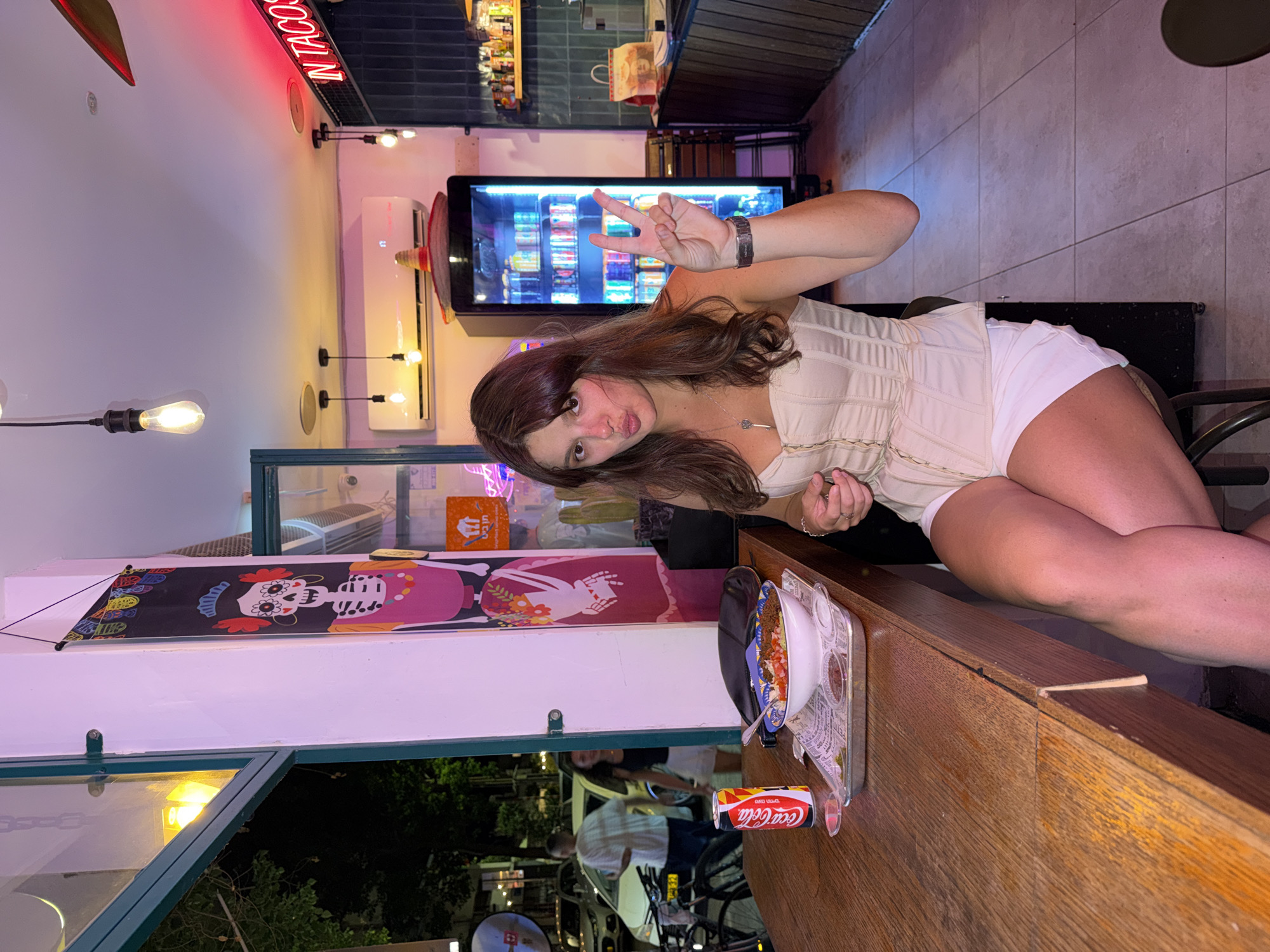 A young woman with brown hair is sitting at a wooden table in a brightly lit restaurant in Tel Aviv, Israel. She is wearing a beige corset top and white shorts, and she is holding up two fingers in a peace sign. In front of her on the table is a bowl of food, a can of Coca-Cola, and some small containers of sauce. Behind her, a refrigerator is stocked with drinks, and a neon sign that reads "IN TACOS" is visible on the wall. The restaurant appears to be decorated with Mexican-themed items, such as a skeleton banner and a sombrero. The overall atmosphere is casual and lively.