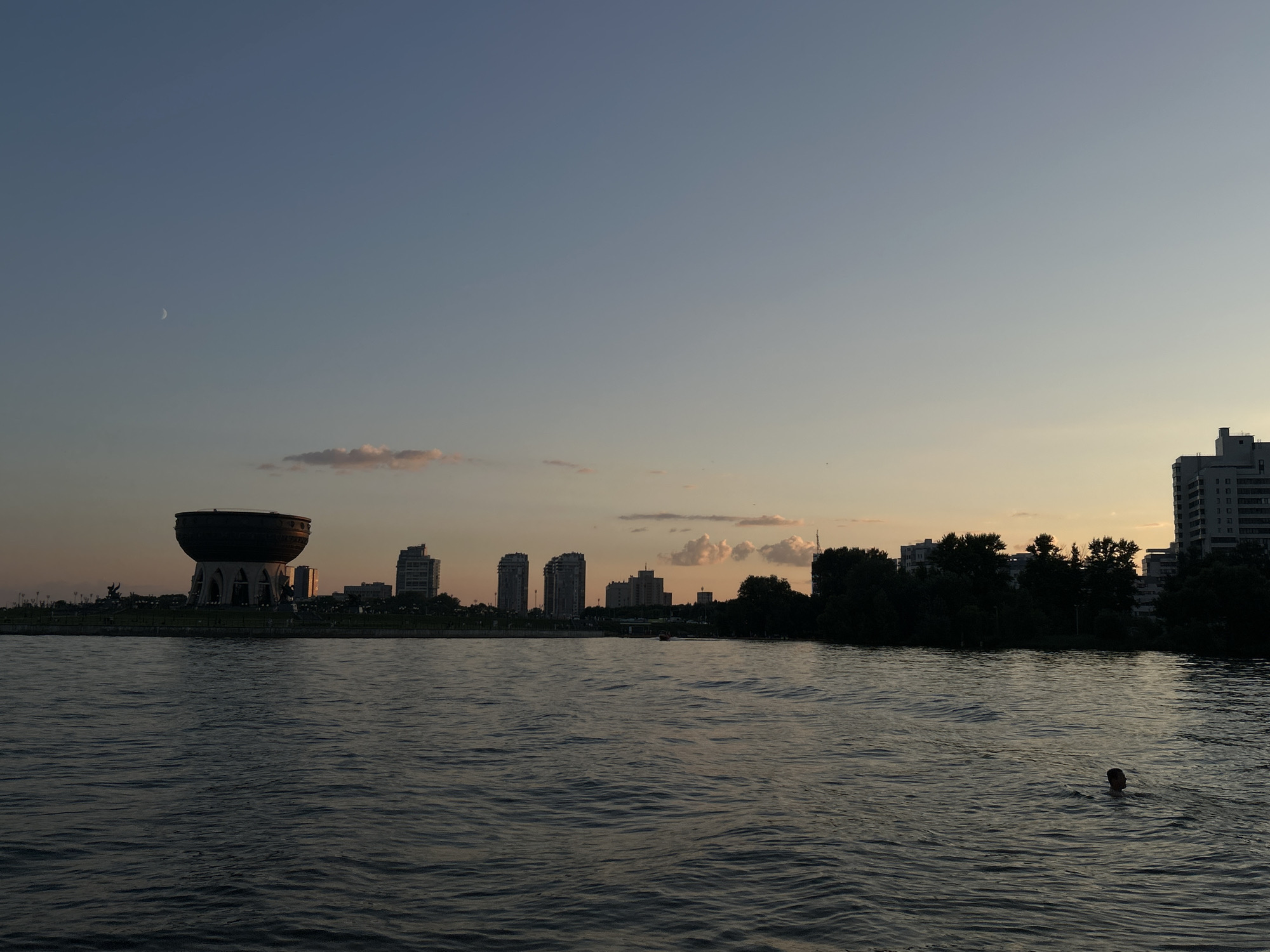 This is a dusk photograph of the Kazanka River in Kazan, Russia, with the city skyline visible in the distance. On the left side of the frame, a distinctive bowl-shaped building with ornate supports, likely the "Family Center 'Kazan'", stands prominently. Several modern high-rise buildings are visible to its right, along with a dense line of trees forming the riverbank. The sky is a gradient of light blue and pale orange, with a sliver of the crescent moon visible near the top left. The water of the river is rippled, reflecting the muted twilight colors. In the lower right quadrant of the image, a single person is partially submerged in the water, with only their head and shoulders visible. The overall impression is a serene urban landscape at the transition from day to night.