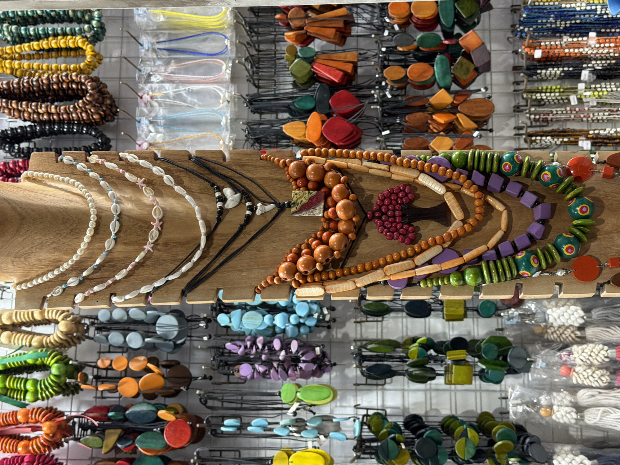 This is a medium-angle, eye-level, outdoor shot of a vibrant display of handmade jewelry in Kuta, Indonesia. The scene is brightly lit, suggesting daytime. The jewelry is predominantly made of natural materials like wood and shells, and features a wide array of colors including greens, blues, purples, oranges, reds, and natural wood tones.
The subjects are the many strands of necklaces and bracelets, meticulously arranged on wooden stands and metal racks. The necklaces vary greatly in style, from delicate strings of small white pearls and shells to chunky, colorful wooden beads. Some necklaces feature unique pendants or charms. The bracelets are also diverse, with some appearing as thick bands of similarly colored beads and others as more intricate arrangements.
The background consists of a wire grid system, typical of market stalls, displaying more jewelry. To the right, several packages containing white, rope-like items are visible, possibly another type of accessory or craft material. The overall impression is that of a bustling marketplace, likely a souvenir shop or artisan stall, brimming with colorful and diverse handmade items. There is no visible text in the image.