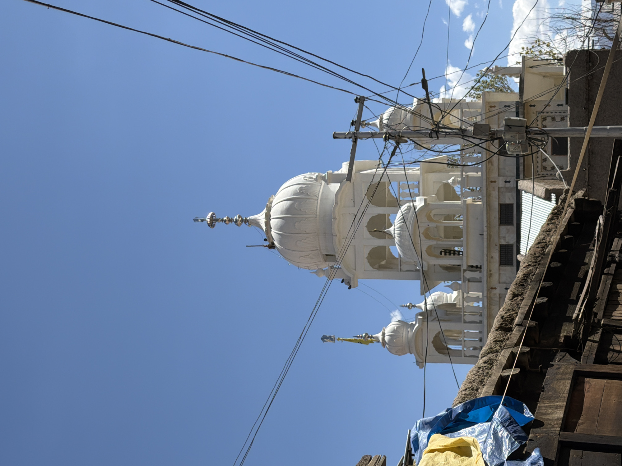 The image captures the exterior of a Sikh gurdwara, likely in the city of Leh, India, under a clear blue sky. The central subject is a white, multi-domed building with intricate architectural details, including arched windows and decorative finials. In the foreground, there's a lower, older structure with a thatched roof, partially covered by blue and yellow tarps. Several power lines crisscross the frame, obscuring parts of the building and the sky. A utility pole with numerous wires is visible on the right side of the image. The scene appears to be a blend of religious architecture and the everyday urban environment, characterized by the presence of power lines and weathered structures.