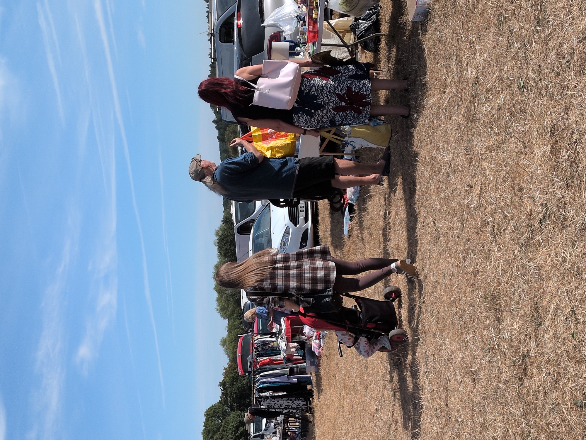 This image depicts an outdoor scene, likely a car boot sale or market, on a sunny day in Claverley, United Kingdom. The ground is covered in dry, light brown grass. In the foreground, a woman with long brown hair is pushing a stroller. The stroller contains a child wearing a light-colored outfit with red fruit patterns. The woman is wearing a brown and white plaid tunic over dark leggings and white and brown sneakers.
Further back, a man with long blond hair and a baseball cap is standing, wearing a blue t-shirt and dark shorts. He is holding a yellow shopping bag and appears to be interacting with a woman with long red hair. This woman is wearing a black sleeveless top and a patterned skirt with a blue, white, and red design. She is carrying a light pink tote bag over her right shoulder and a yellow and white shopping bag in her left hand.
In the background, there are several vehicles, including white cars and a white van. Clothes racks are set up with various items of clothing for sale. Tables are laden with goods, and other people can be seen browsing. The sky is a clear blue with a few wispy white clouds. Trees are visible in the distance, suggesting a rural or semi-rural setting. The overall impression is one of a casual community event.