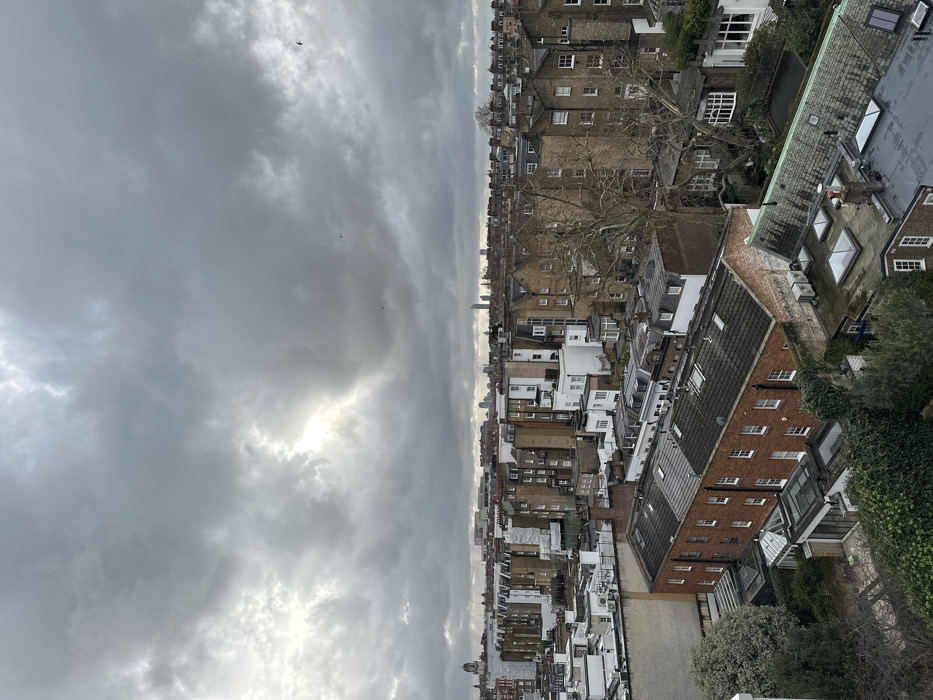 This image presents an elevated, wide-angle view of a densely packed urban residential area in London, United Kingdom, specifically looking eastward across a section of South Kensington or its adjacent districts. The sky is extensively covered by heavy, dark grey stratocumulus clouds, with a distinct brighter patch of diffused sunlight visible towards the west-northwest. Several small, dark avian subjects are in flight across the upper portion of the sky.
The scene is dominated by a vast expanse of traditional London architecture, primarily comprising multi-story terraced and semi-detached houses. These structures predominantly feature brick facades, some with painted stucco, and exhibit a variety of pitched roofs made from slate and tile. Numerous chimneys, rooftop ventilation units, and skylights are visible across the rooftops, particularly in the foreground. In the mid-ground, one building is partially obscured by white scaffolding and sheeting, indicating ongoing construction or renovation. Interspersed among the buildings are numerous deciduous trees, bare of leaves, characteristic of late autumn or winter, along with patches of green evergreen foliage in private gardens and courtyards below.
On the far-left horizon, a prominent domed building with an adjoining tower structure is identifiable as the Victoria and Albert Museum. Further to the right in the distant background, a cluster of modern, slender high-rise buildings is discernible, marking the skyline of London's central financial districts (e.g., the City of London or Canary Wharf). No human subjects, specific actions, or direct interactions are visible within the frame. The overall environment conveys an established, closely built-up metropolitan landscape under overcast weather conditions.
